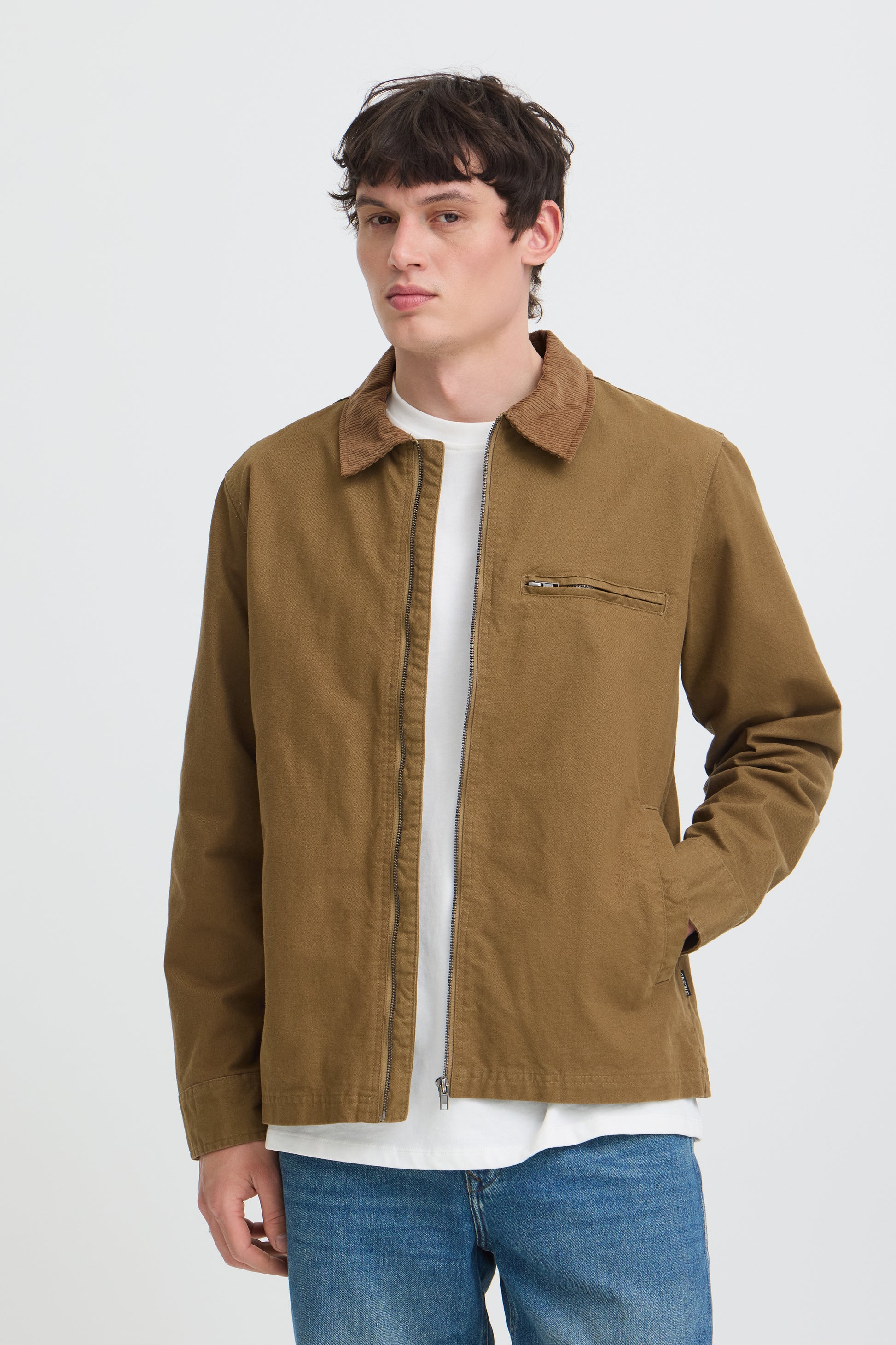 A man wearing a brown utility jacket over a white t-shirt and blue jeans, standing against a light gray studio background.