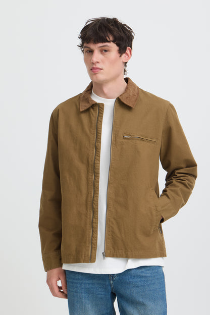 A man wearing a brown utility jacket over a white t-shirt and blue jeans, standing against a light gray studio background.
