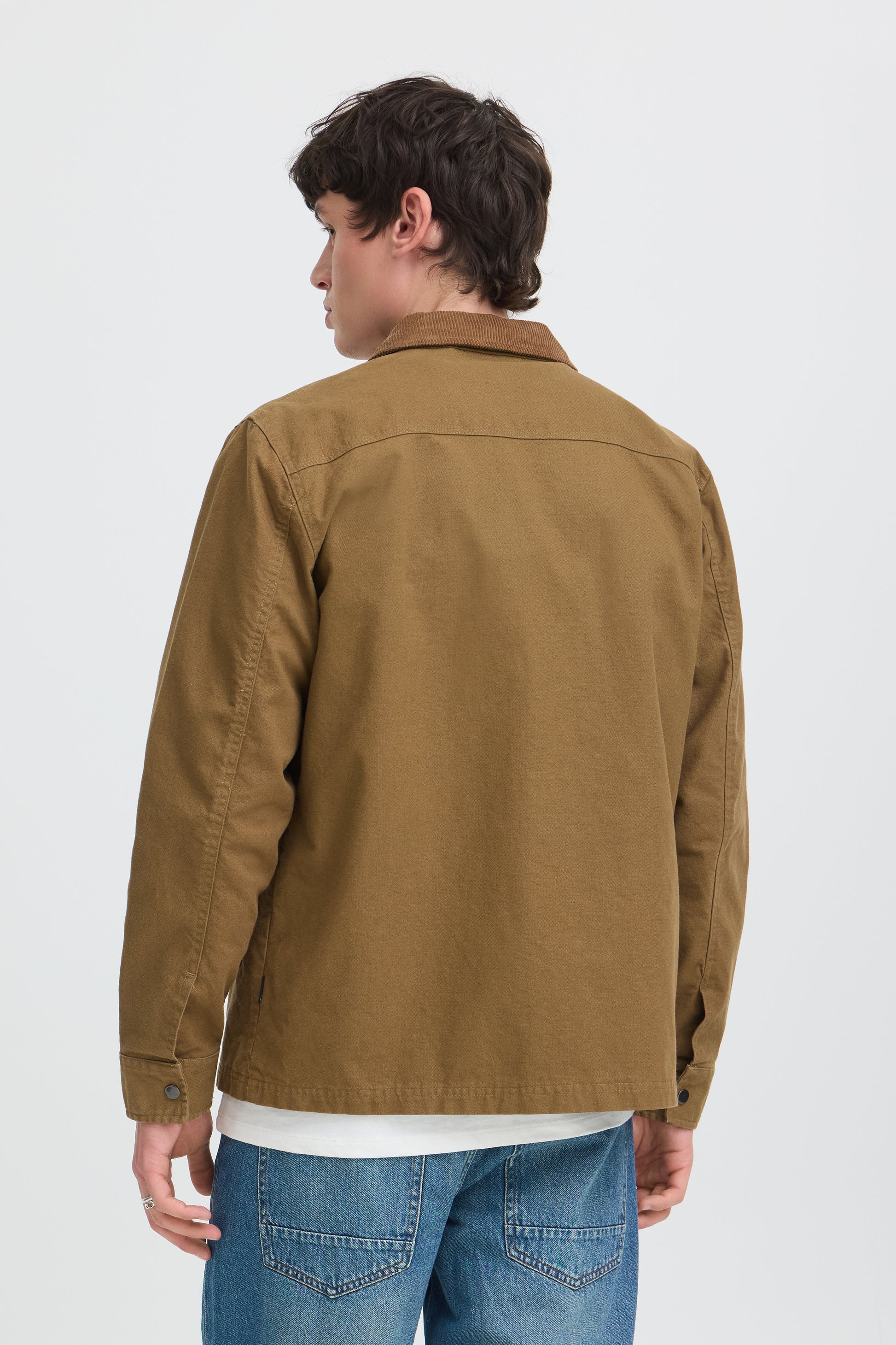 A man shown from the back wearing a brown utility jacket over a white t-shirt and blue jeans, photographed against a light gray studio background.