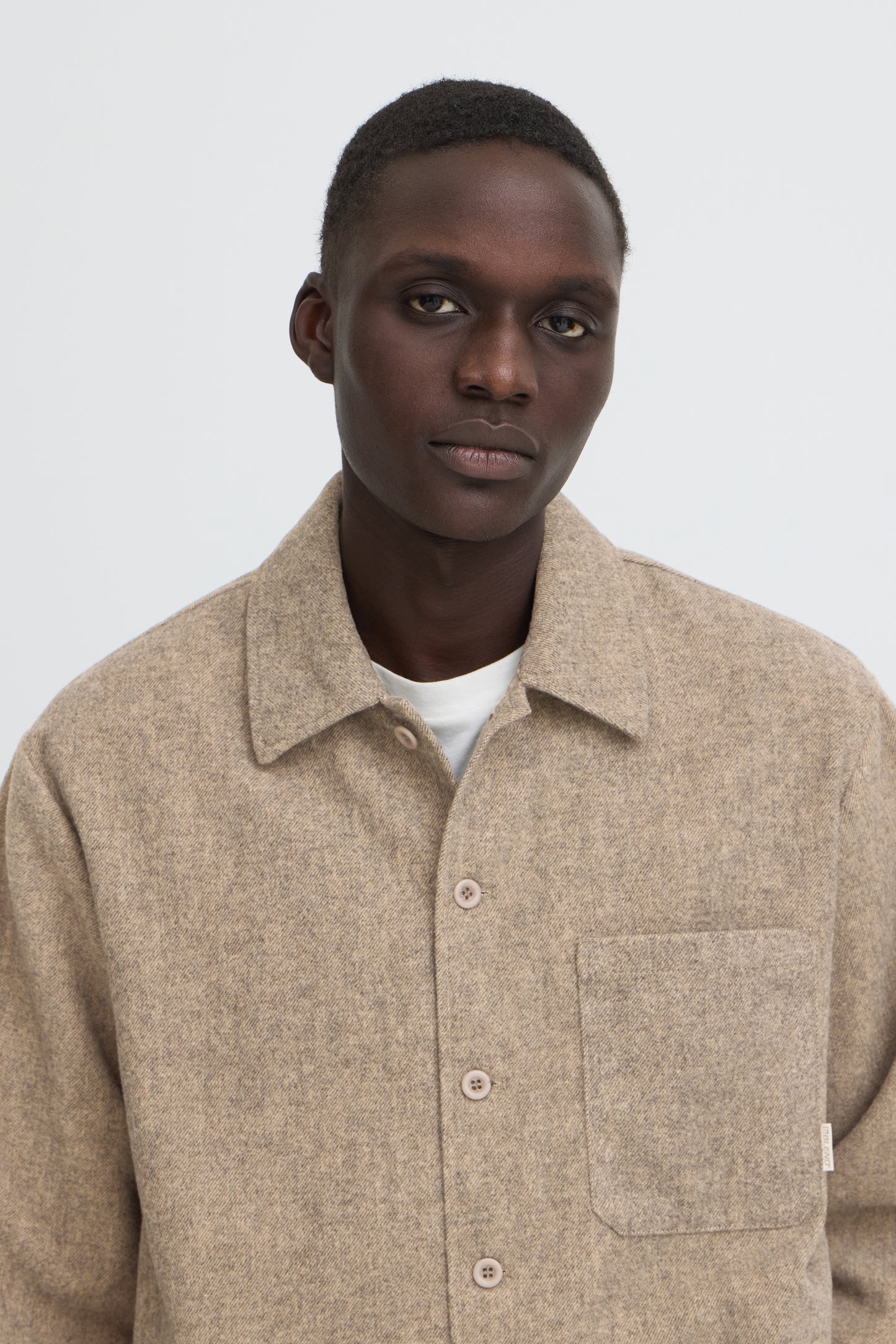A man wearing a light beige textured overshirt with a collar and front buttons over a white t-shirt, photographed against a neutral light gray background.