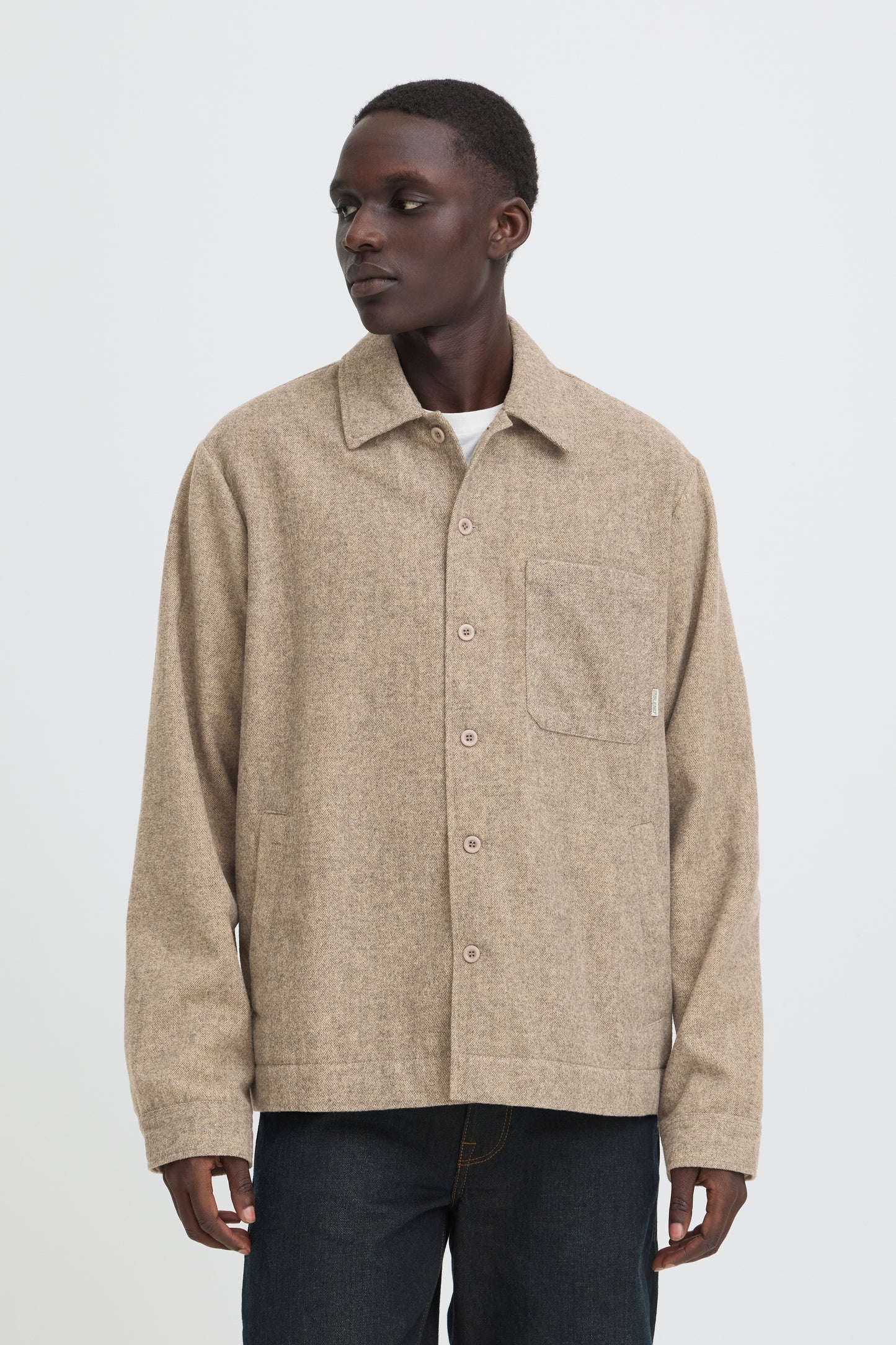 A man wearing a beige textured button-up overshirt with a chest pocket, paired with dark jeans, photographed against a plain light gray studio background.