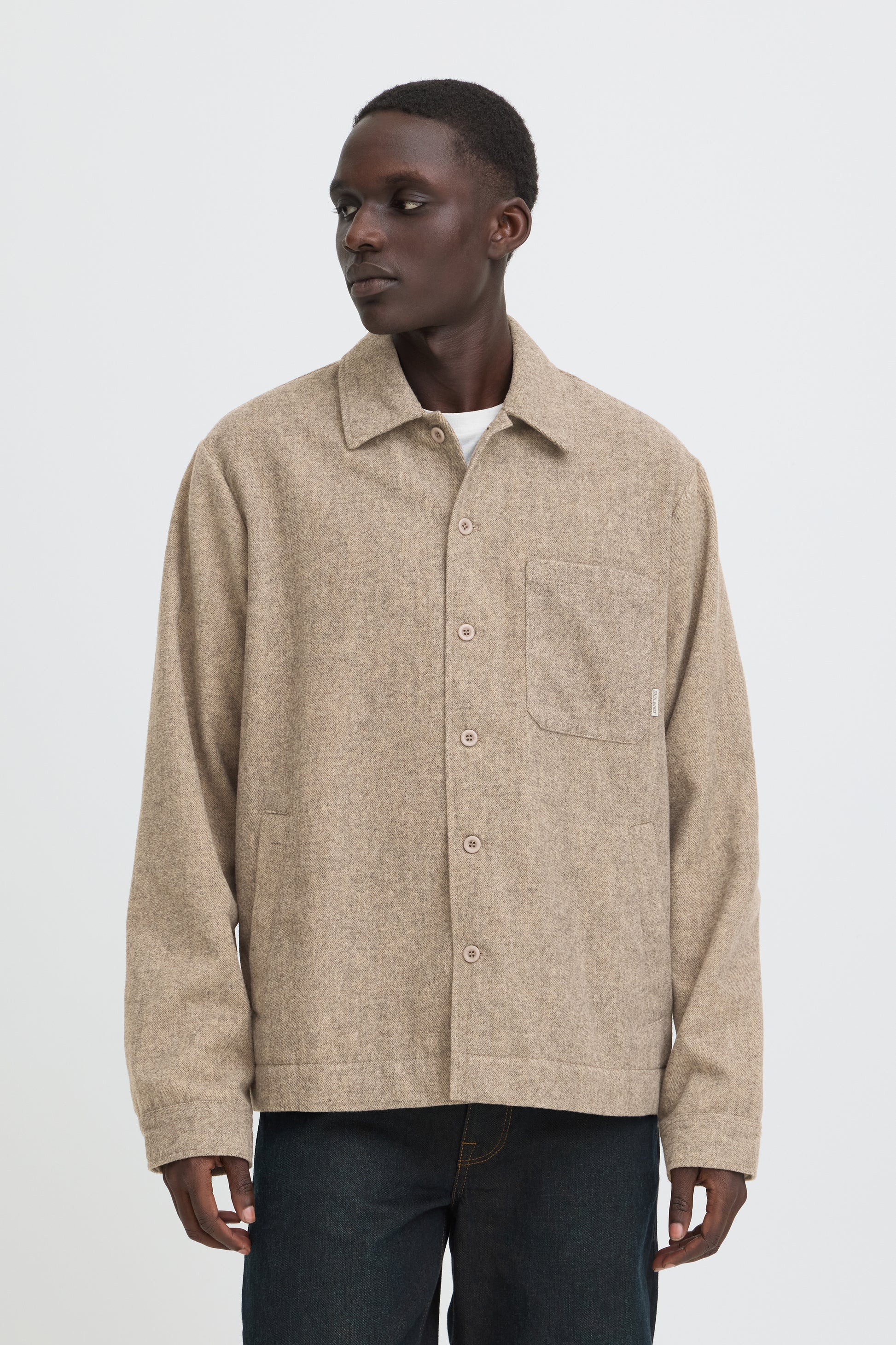 A man wearing a beige textured button-up overshirt with a chest pocket, paired with dark jeans, photographed against a plain light gray studio background.