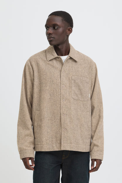 A man wearing a beige textured button-up overshirt with a chest pocket, paired with dark jeans, photographed against a plain light gray studio background.