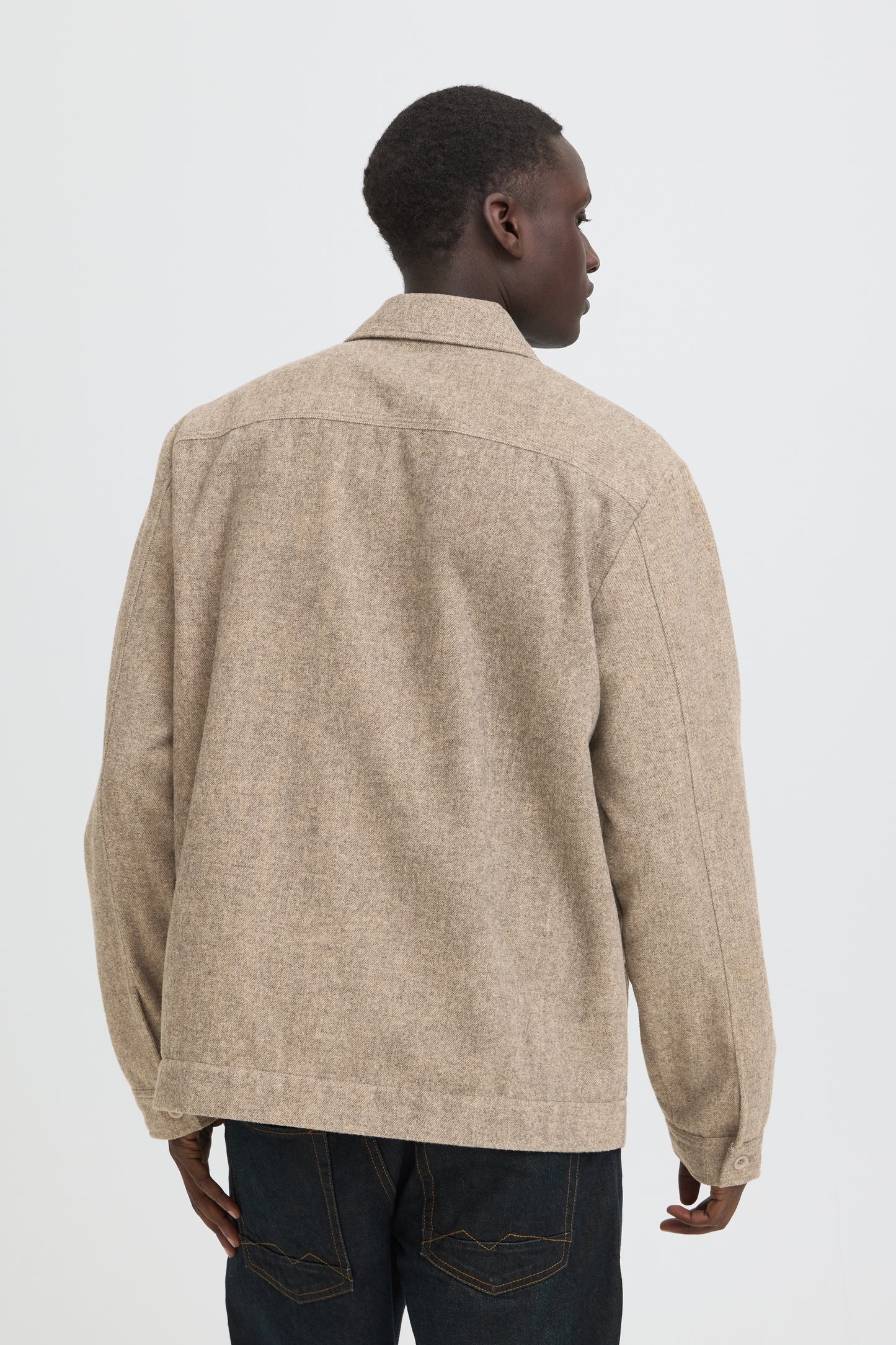 A man viewed from the back wearing a light beige textured overshirt or jacket with dark blue jeans, set against a plain light gray studio background.
