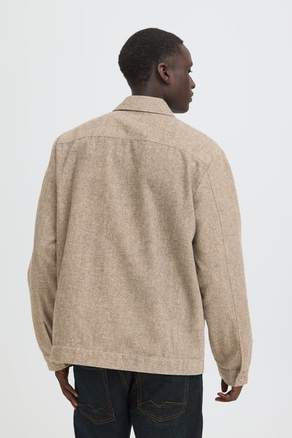 A man viewed from the back wearing a light beige textured overshirt or jacket with dark blue jeans, set against a plain light gray studio background.