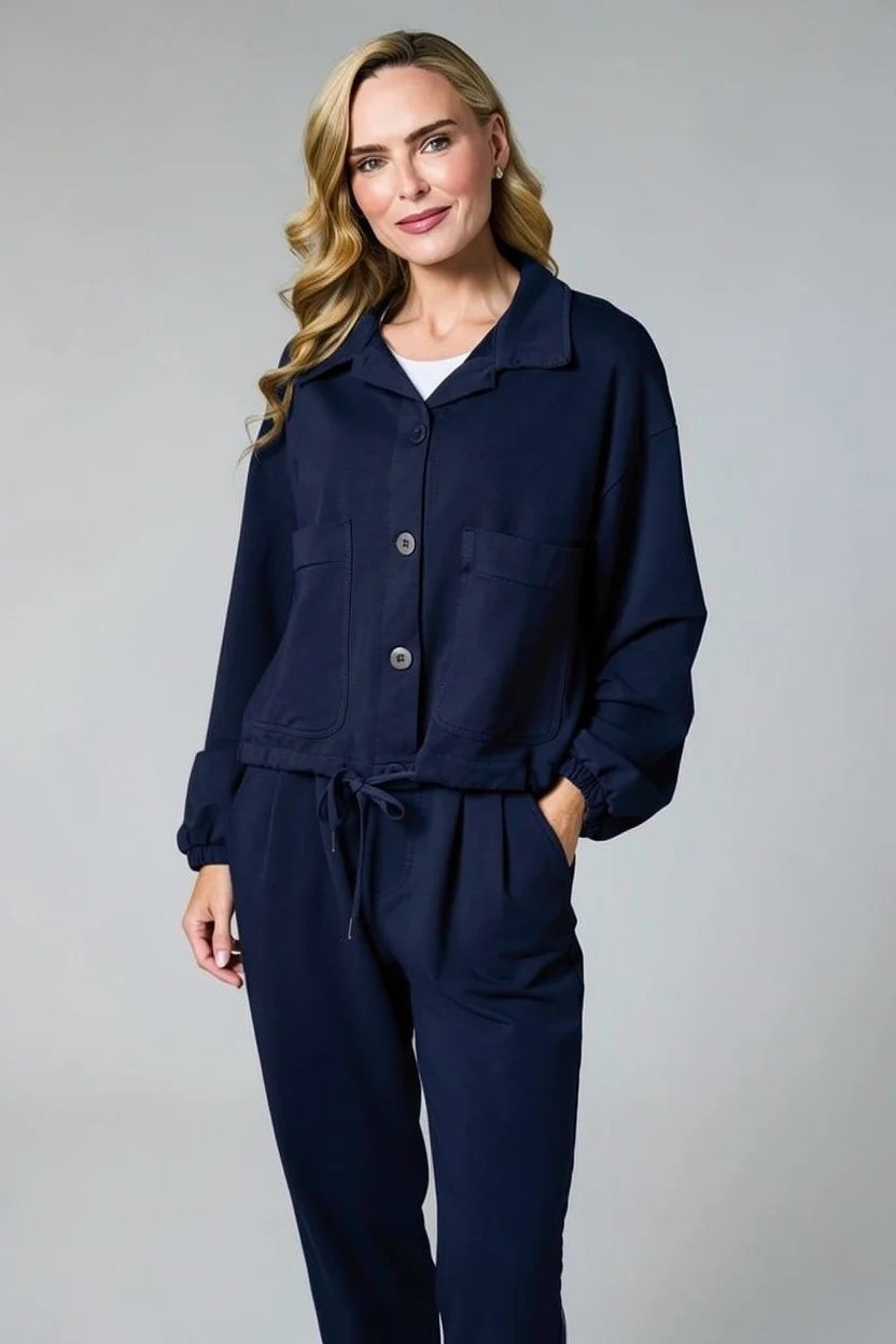 A woman modeling a matching navy blue lounge set consisting of a cropped button-front jacket and coordinating drawstring pants, photographed against a neutral gray studio background.