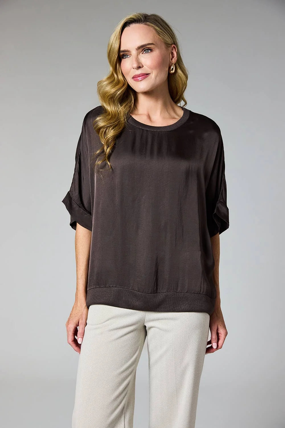 A woman modeling a dark brown short-sleeve top with rolled cuffs and light beige pants against a neutral gray studio background.