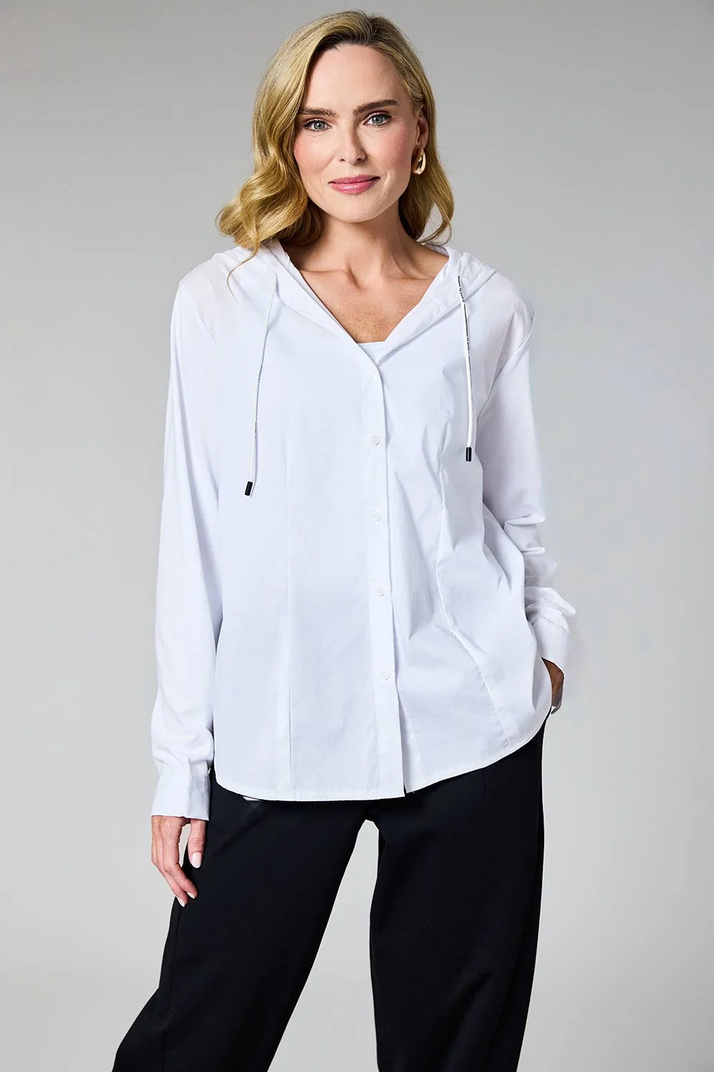 A woman modeling a white long-sleeve hooded button-up shirt with black drawstrings, paired with black pants, against a solid light gray studio background.