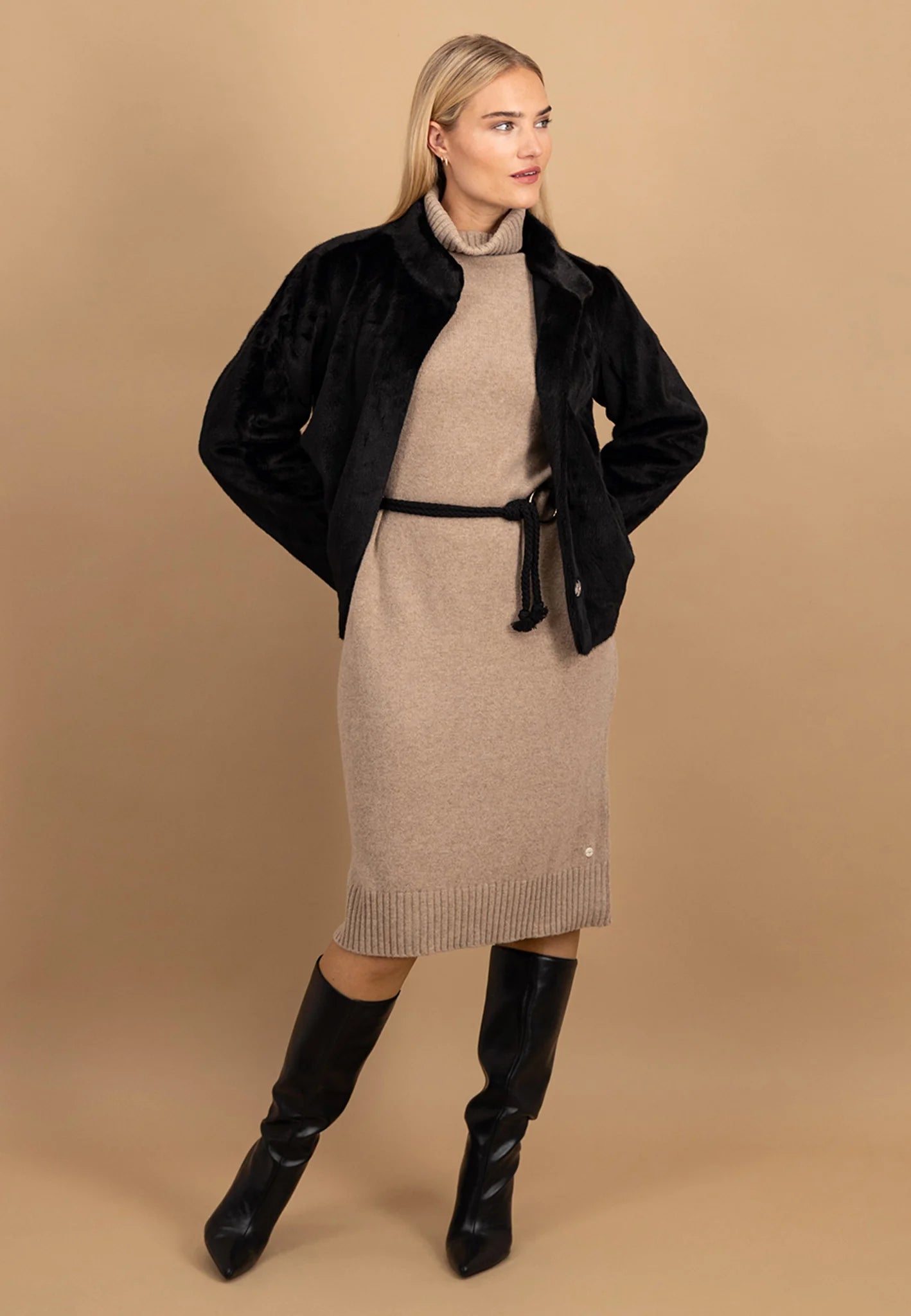Woman wearing a black coat over a beige dress with black boots on a beige background