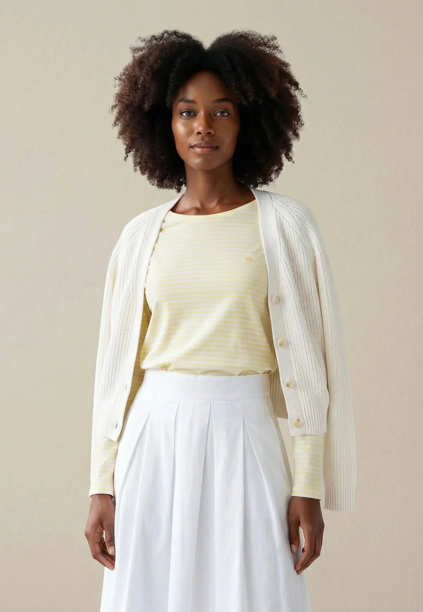 Woman wearing a light yellow top and white skirt with a beige cardigan against a beige background