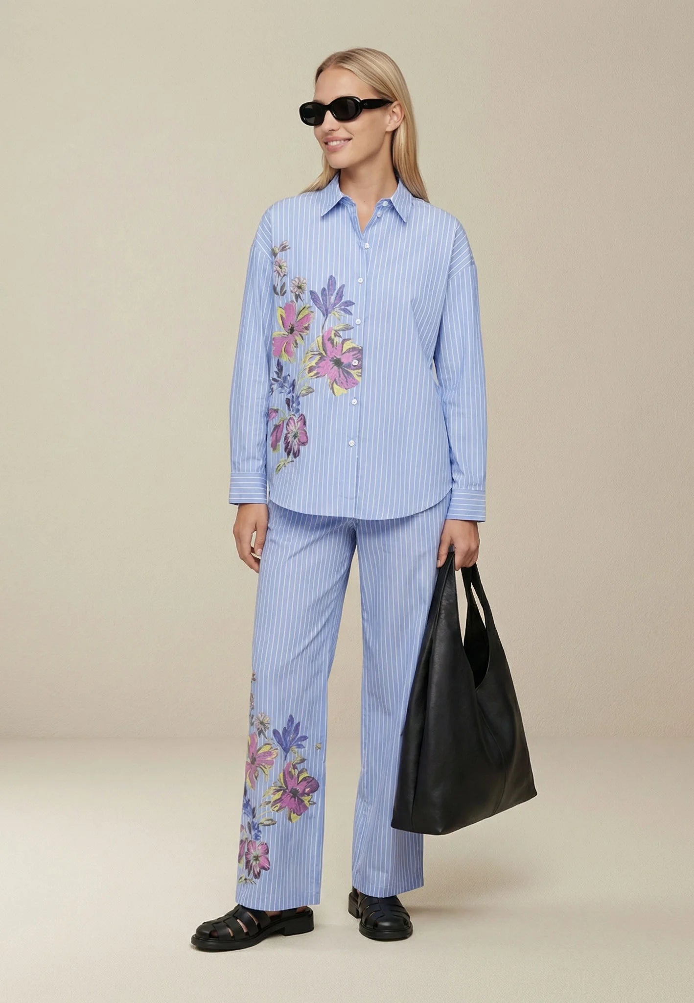 Woman wearing a blue striped outfit with floral embroidery, holding a black bag on a beige background