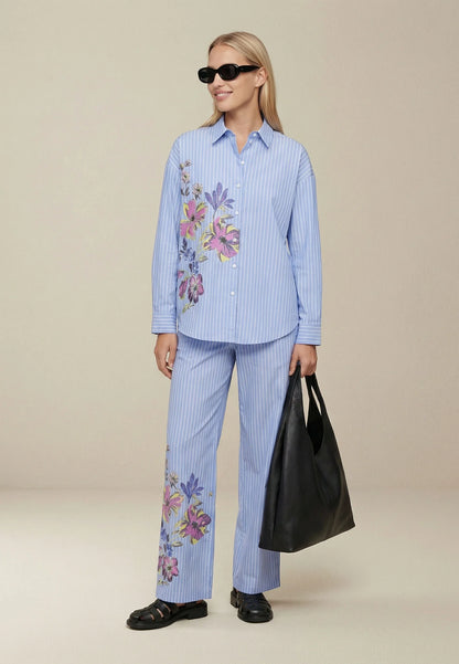 Woman wearing a blue striped outfit with floral embroidery, holding a black bag on a beige background