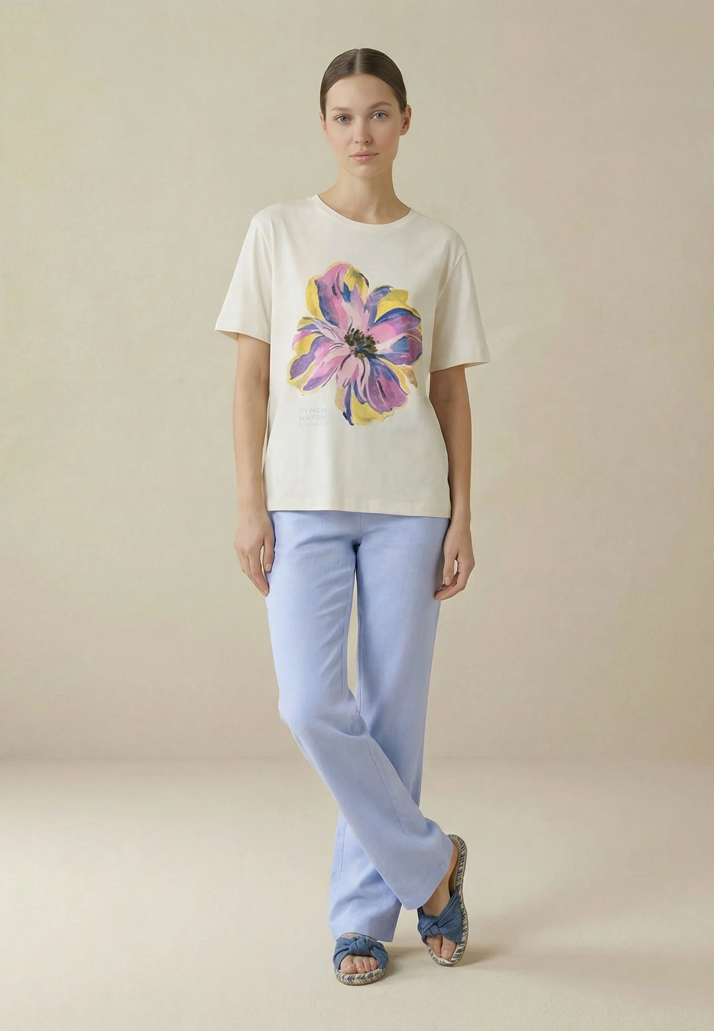 Woman wearing a cream t-shirt with a colorful flower design and light blue pants on a beige background