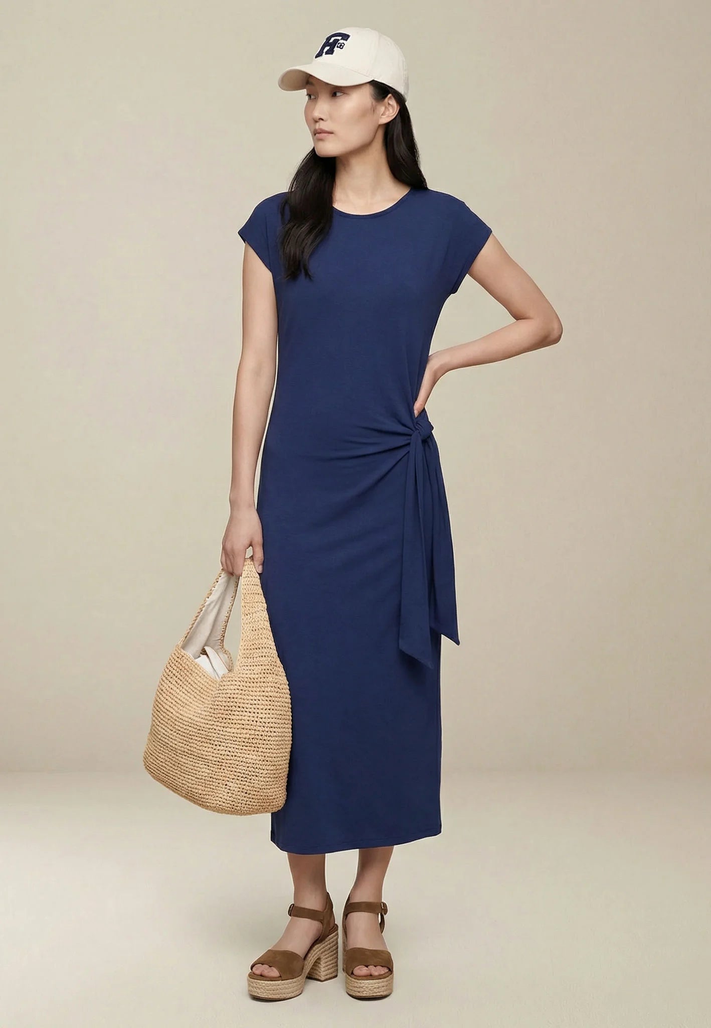 Woman wearing a blue dress and white cap, holding a woven bag on a beige background