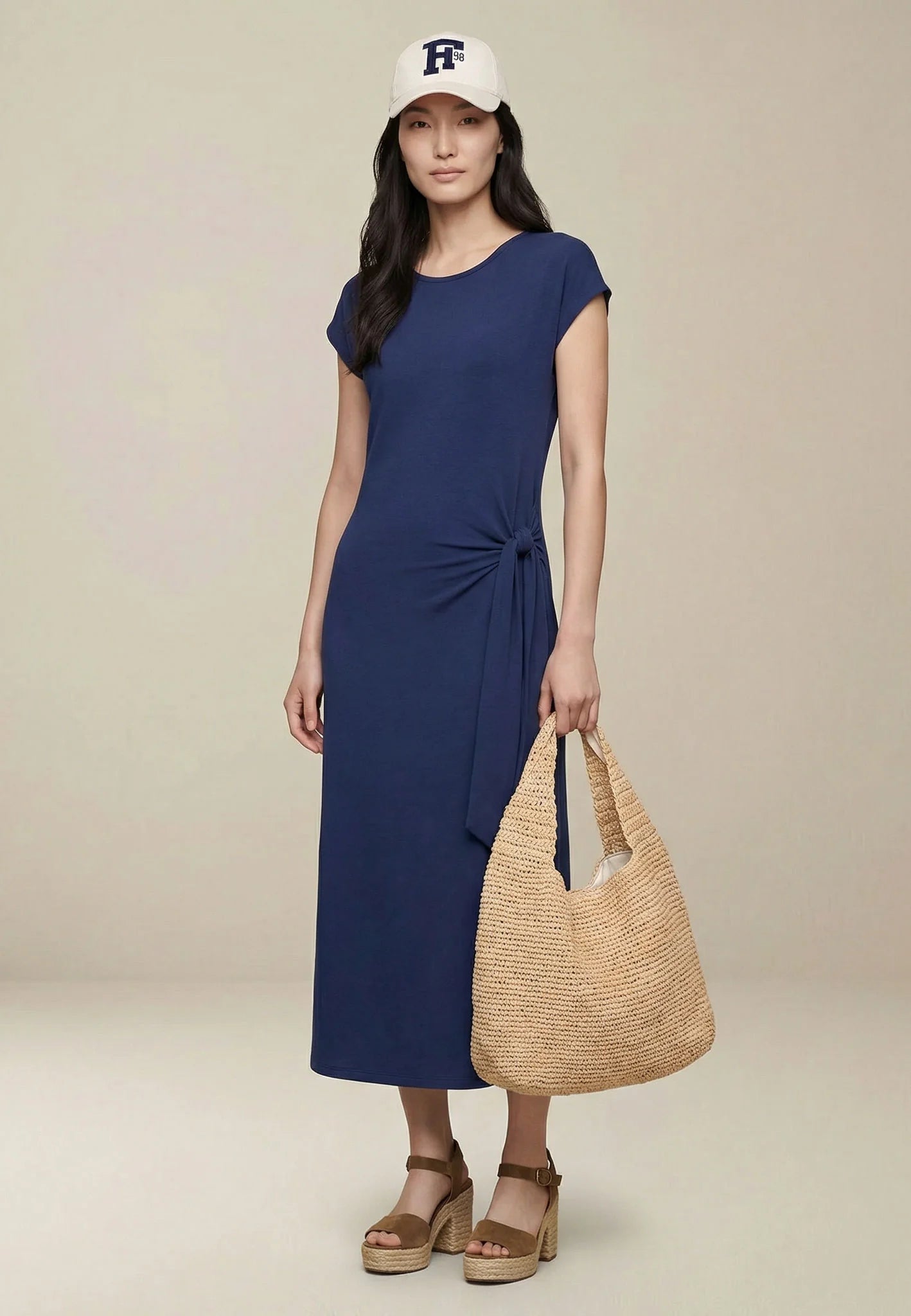 Woman wearing a blue dress and white cap, holding a woven bag on a beige background