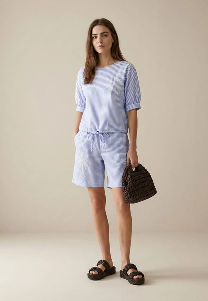 Woman wearing a blue striped outfit with shorts and sandals, holding a woven bag against a beige background.