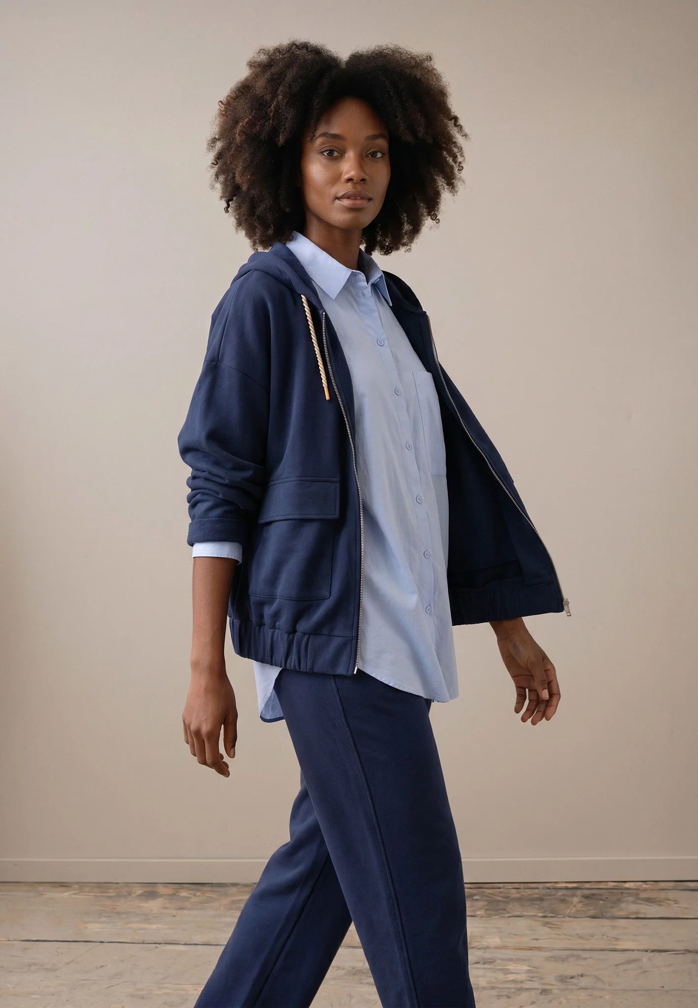 A woman modeling a navy blue zip-up hooded jacket over a light blue button-up shirt and navy trousers, standing against a plain beige studio wall.