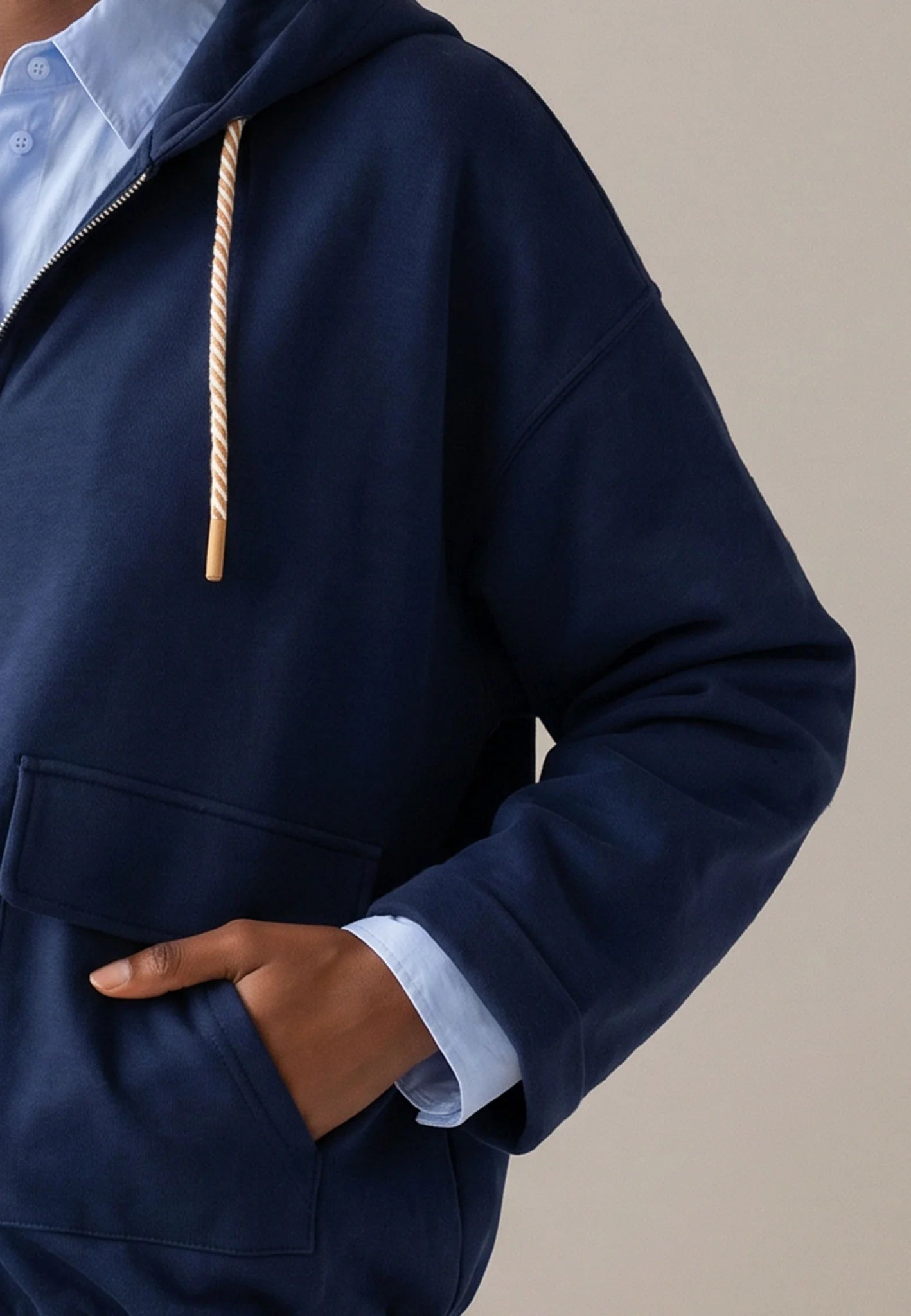 A close-up of a person wearing a navy blue hooded sweatshirt over a light blue collared shirt, with one hand in the front pocket. The hoodie has a cream-colored rope drawstring and is photographed against a plain beige background.