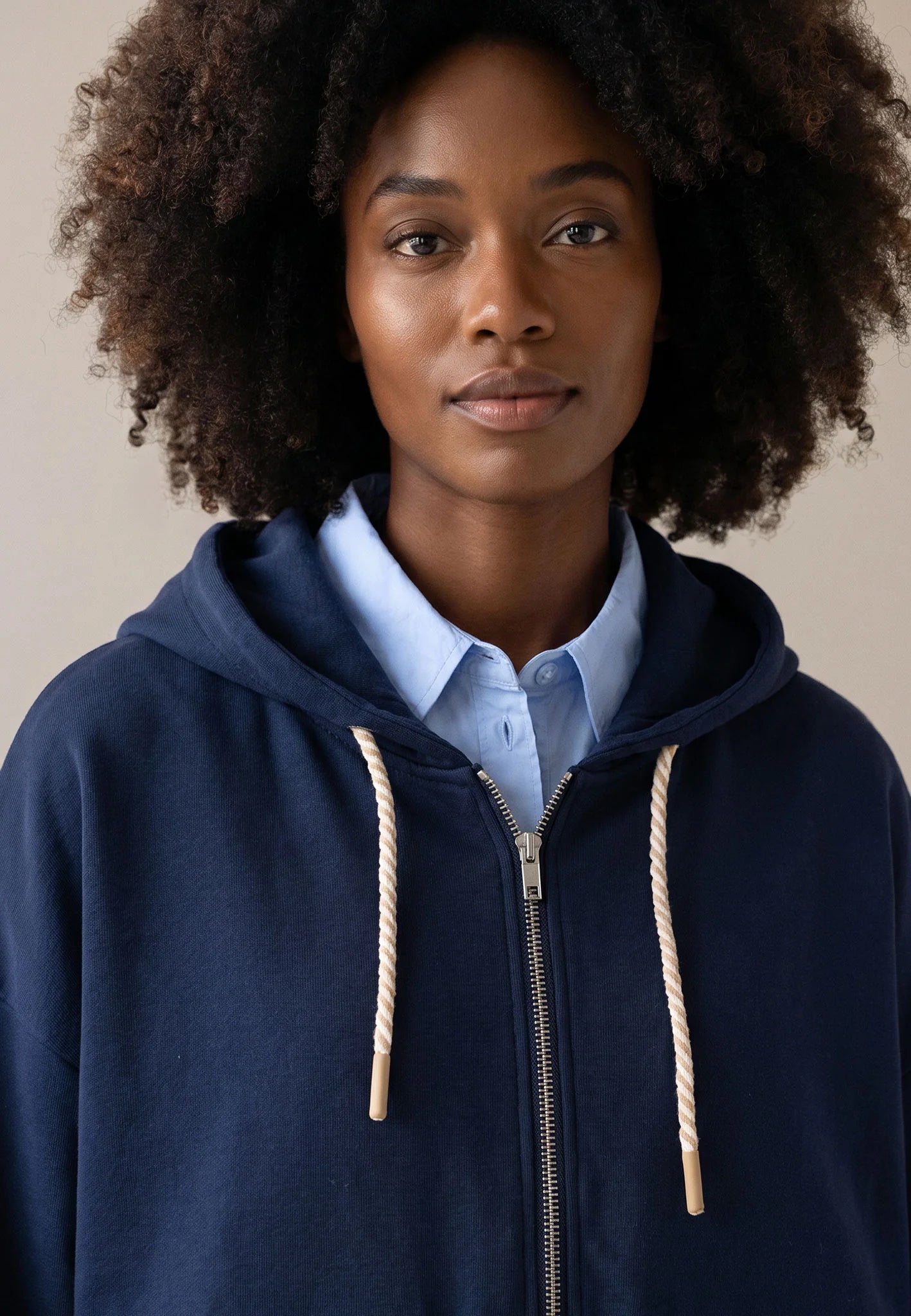 A woman with dark curly hair wearing a navy blue zip-up hoodie over a light blue collared shirt, photographed against a plain beige background.