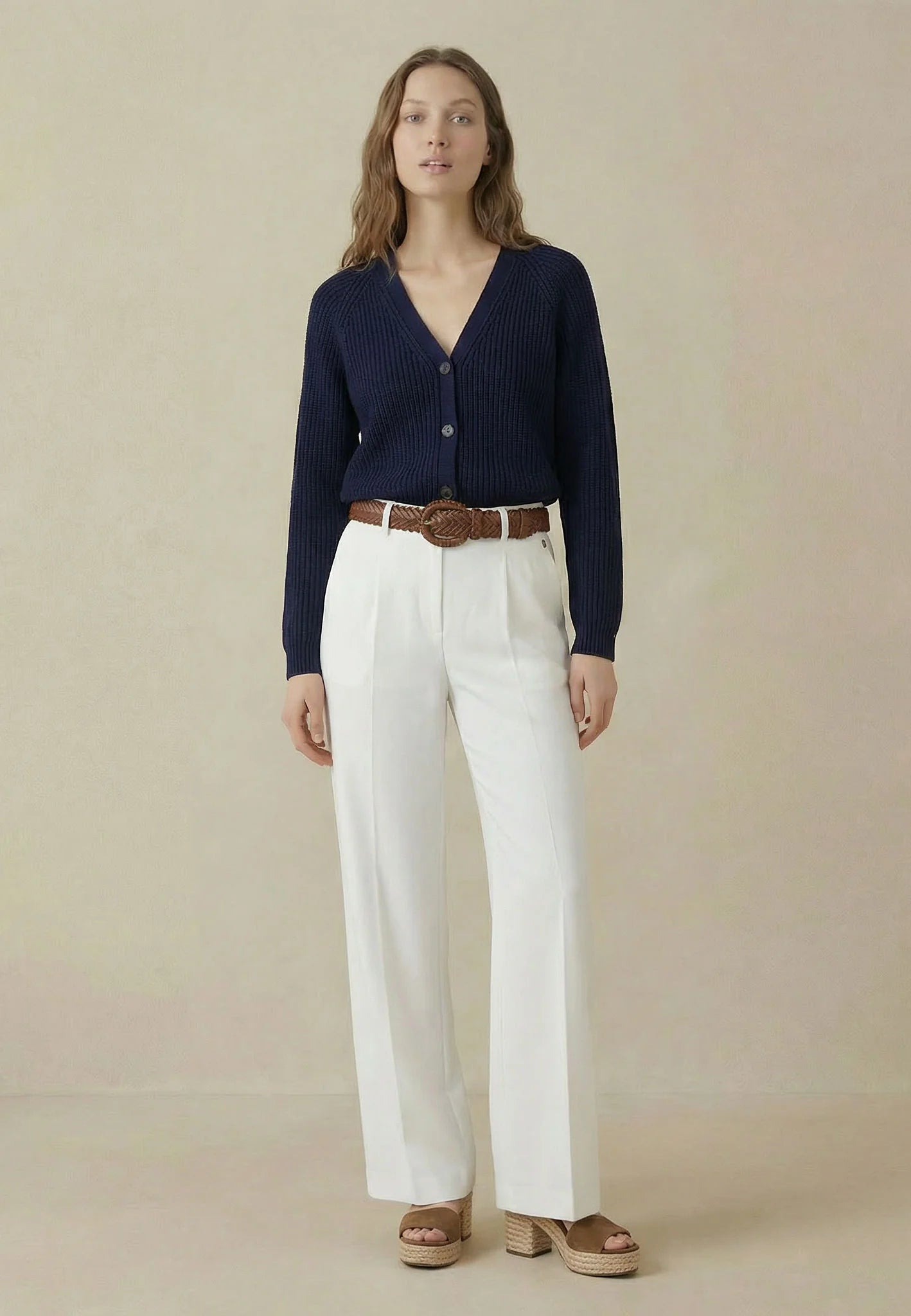 Woman wearing a navy cardigan, white pants, and brown belt against a beige background