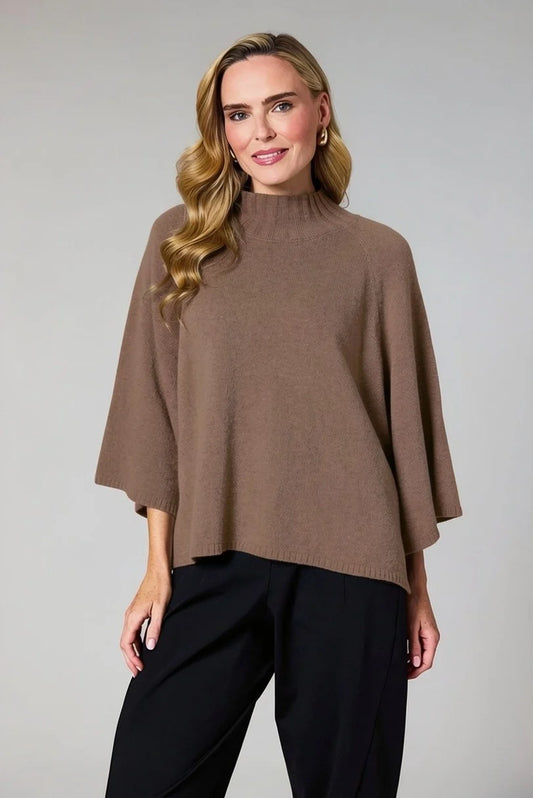 A woman modeling a taupe brown knit poncho sweater with a mock neck and wide draped sleeves, paired with black trousers, against a solid light gray studio background.