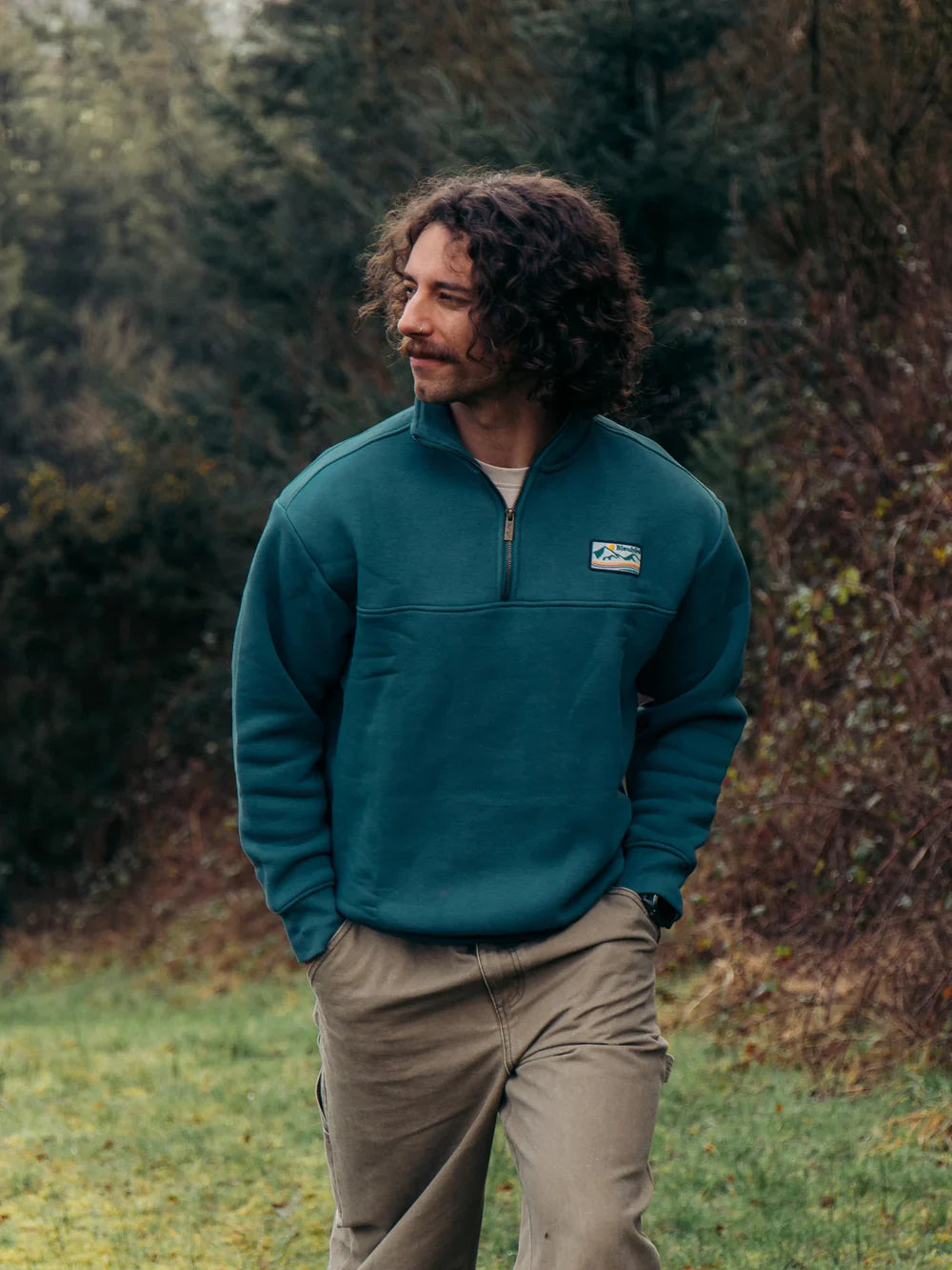 Man wearing a teal pullover with a logo standing in a forested area