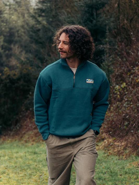 Man wearing a teal pullover with a logo standing in a forested area