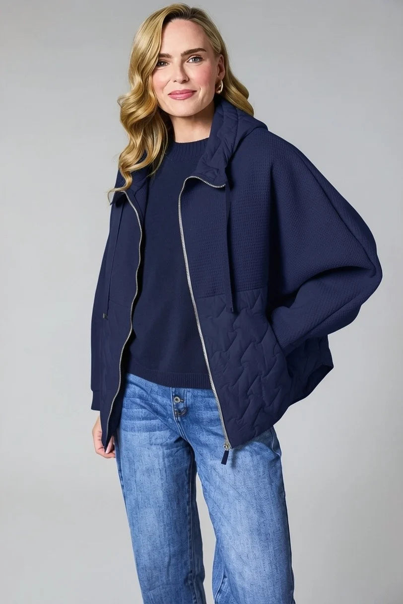 A woman modeling a navy blue quilted hooded jacket with a silver zipper, worn over a matching navy top and blue jeans, against a solid gray studio background.