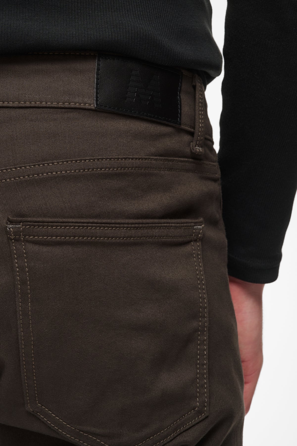 Close-up of the back waistband and pocket of dark brown pants, worn with a black long-sleeve top. A black leather brand patch is visible at the back waistband.