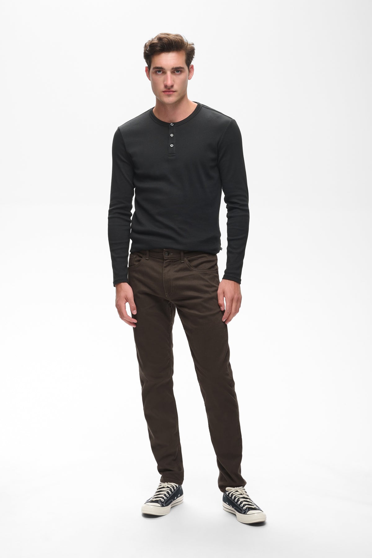 A man modeling a black long-sleeve henley shirt and dark brown slim-fit pants with black and white sneakers, standing against a plain white studio background.