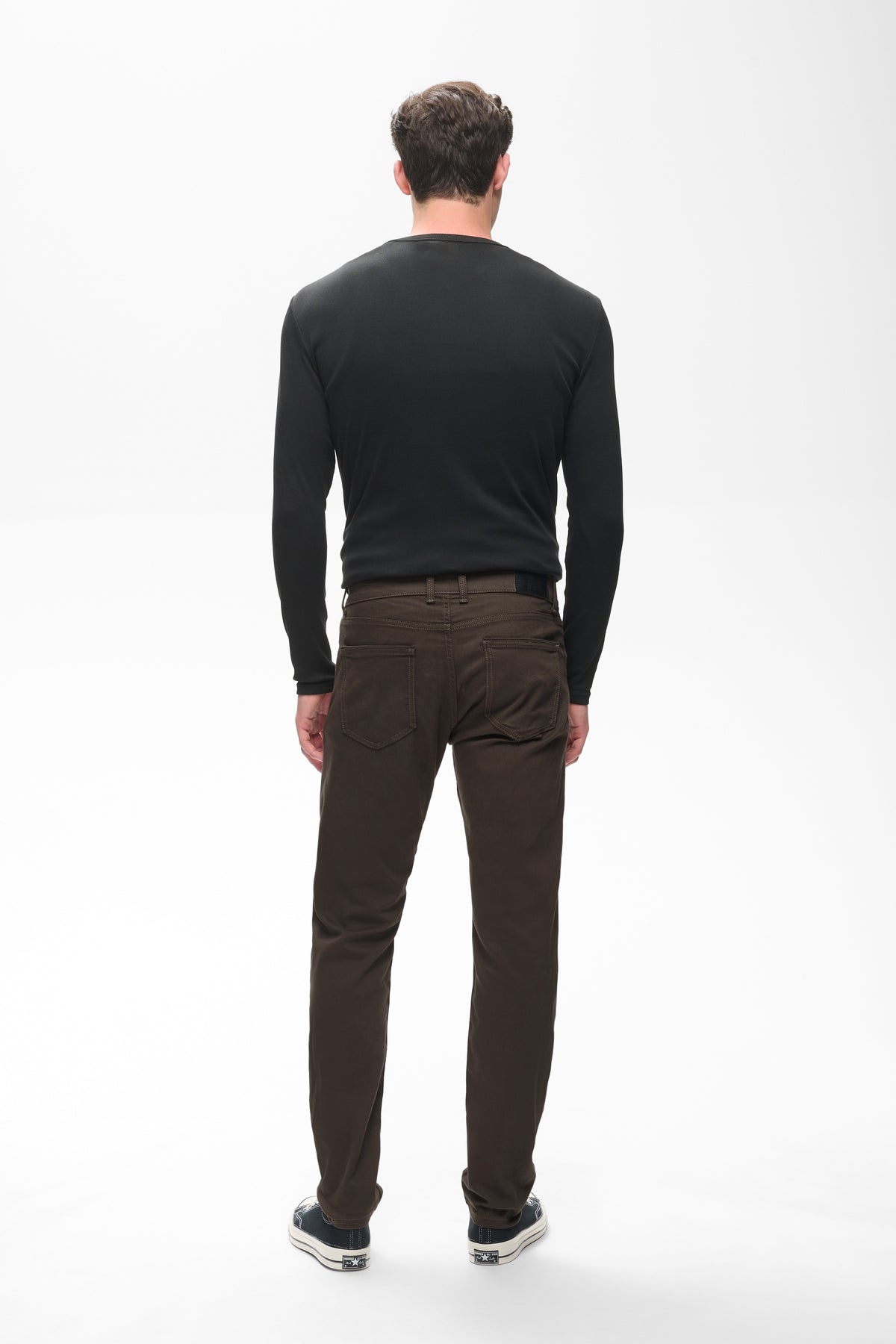 A man viewed from behind wearing a black long-sleeve crewneck shirt and dark brown trousers, standing against a plain white studio background.