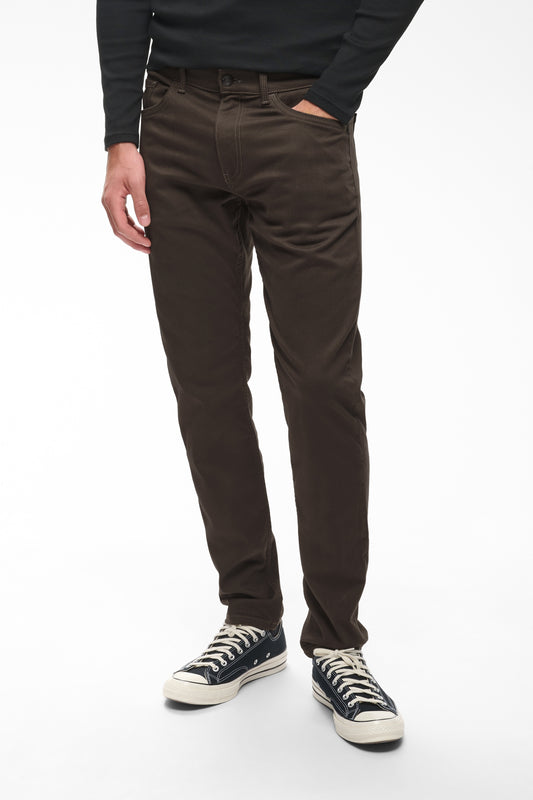 A person wearing slim-fit dark brown chino pants, a black long-sleeve top, and black canvas sneakers with white laces, standing against a plain white studio background.