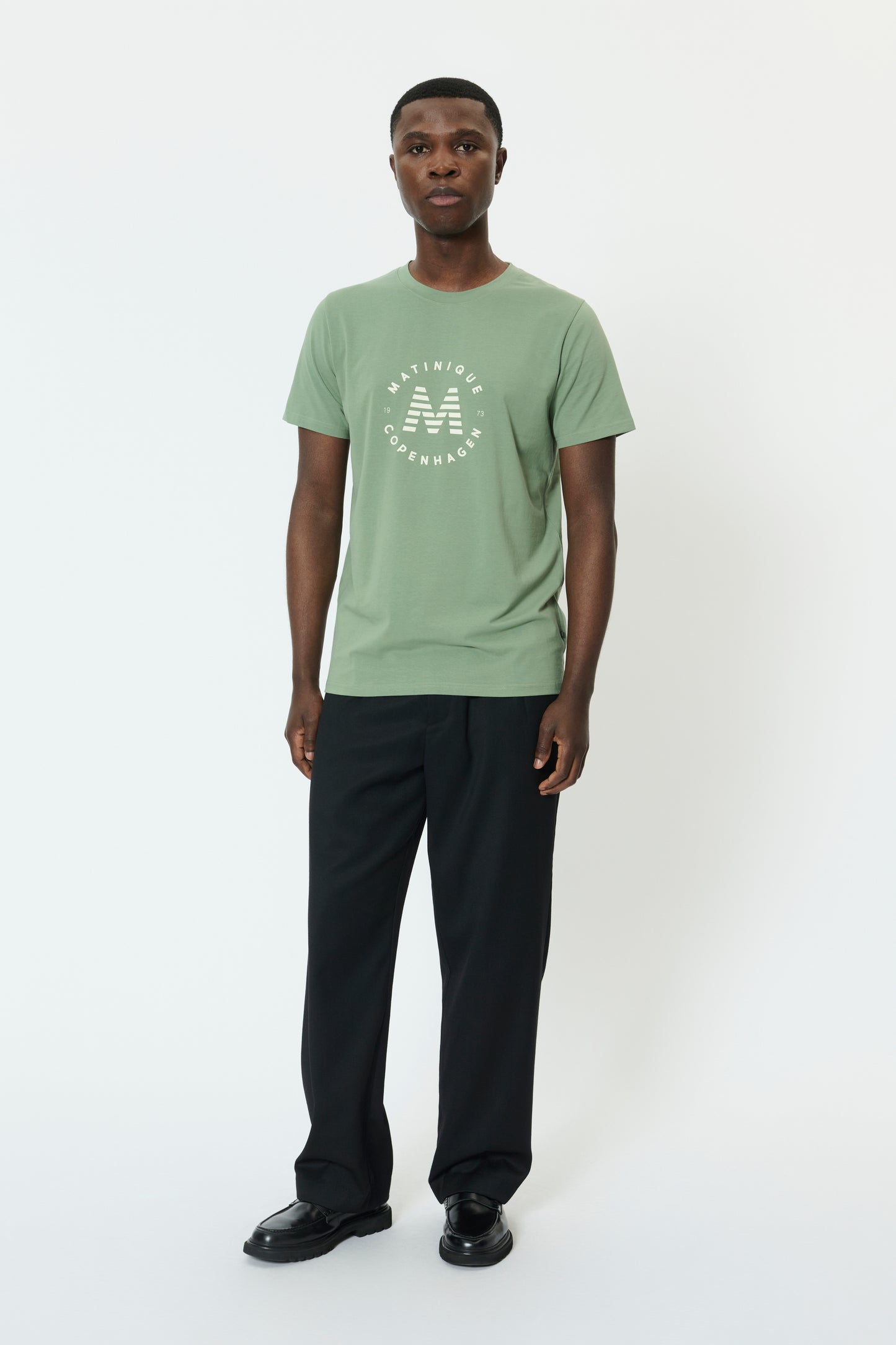 Man wearing a light green t-shirt with a logo and dark pants on a white background