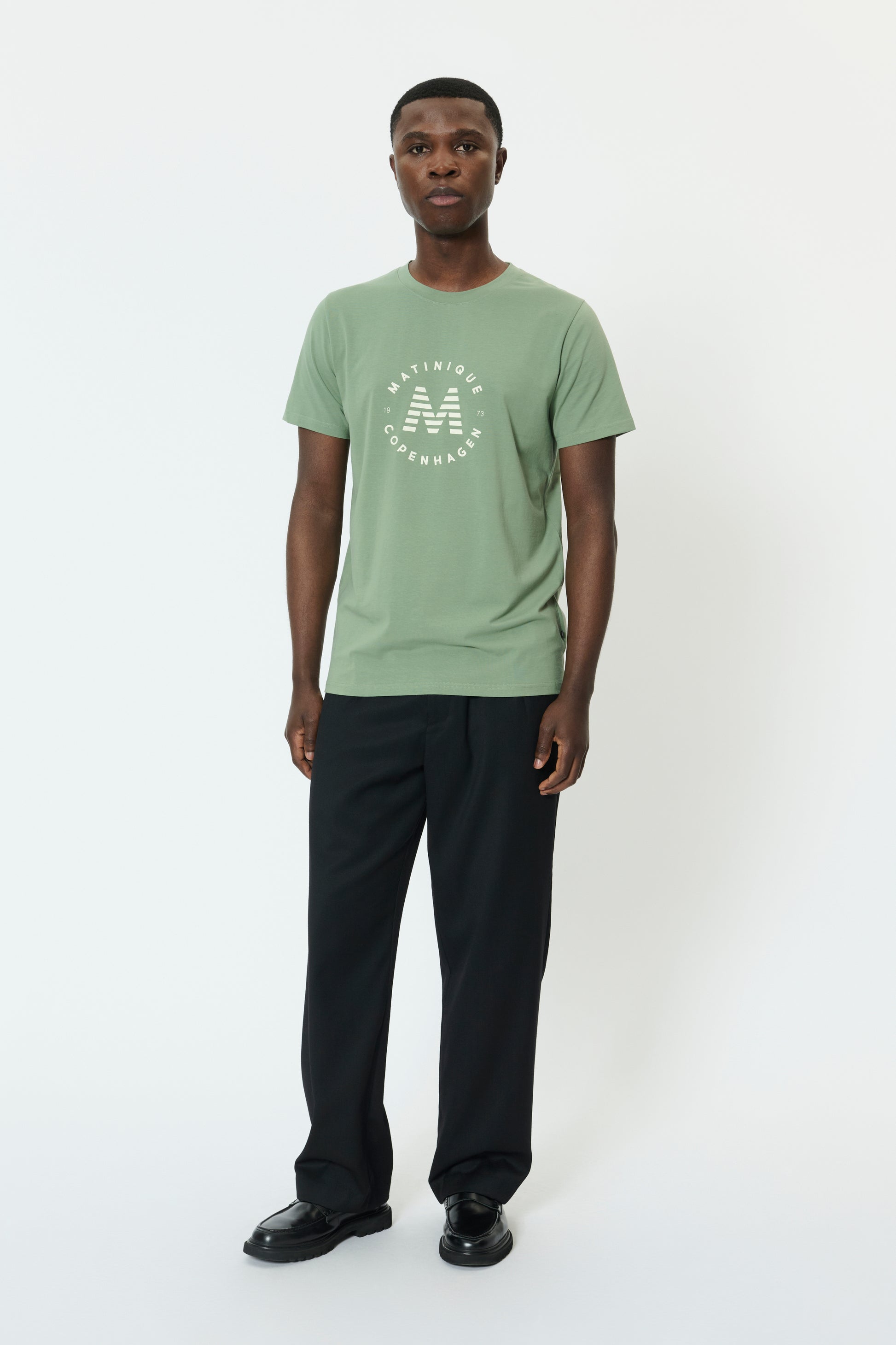 Man wearing a light green t-shirt with a logo and dark pants on a white background
