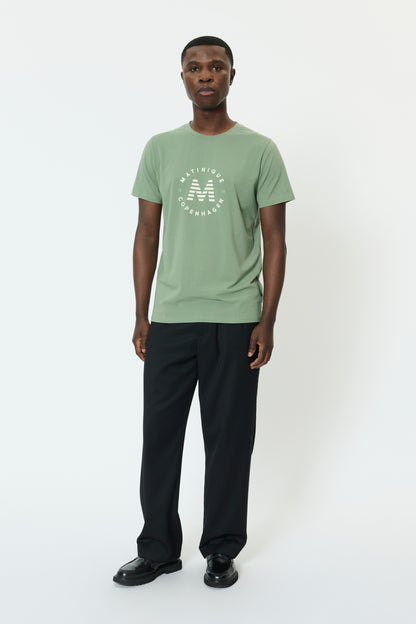 Man wearing a light green t-shirt with a logo and dark pants on a white background