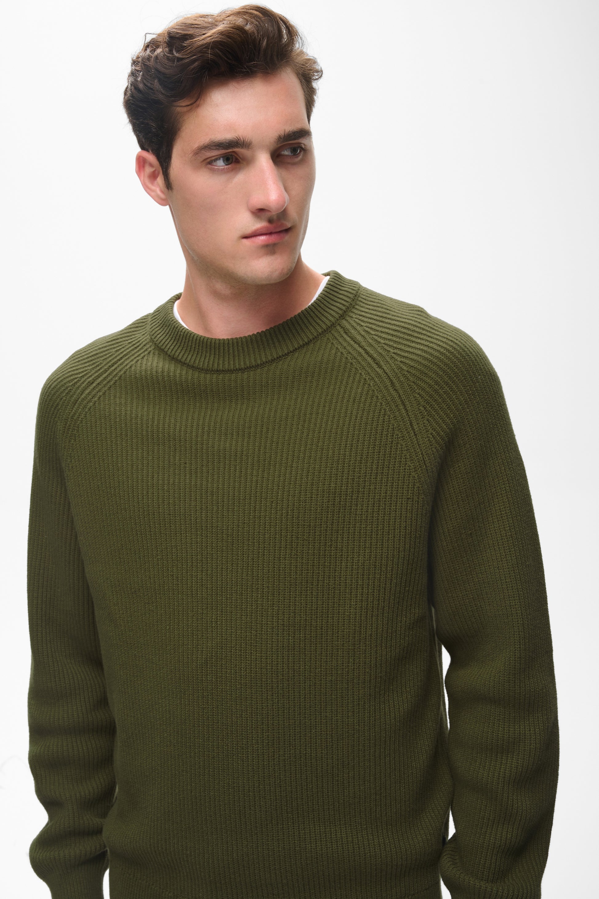 A man wearing a ribbed knit crewneck sweater in olive green, photographed against a plain light gray studio background.