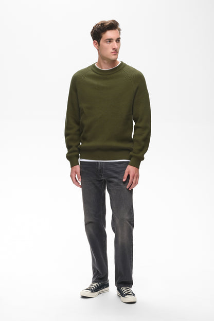 A man modeling a dark olive green crewneck sweater, dark gray jeans, and black and white sneakers against a plain white studio background.