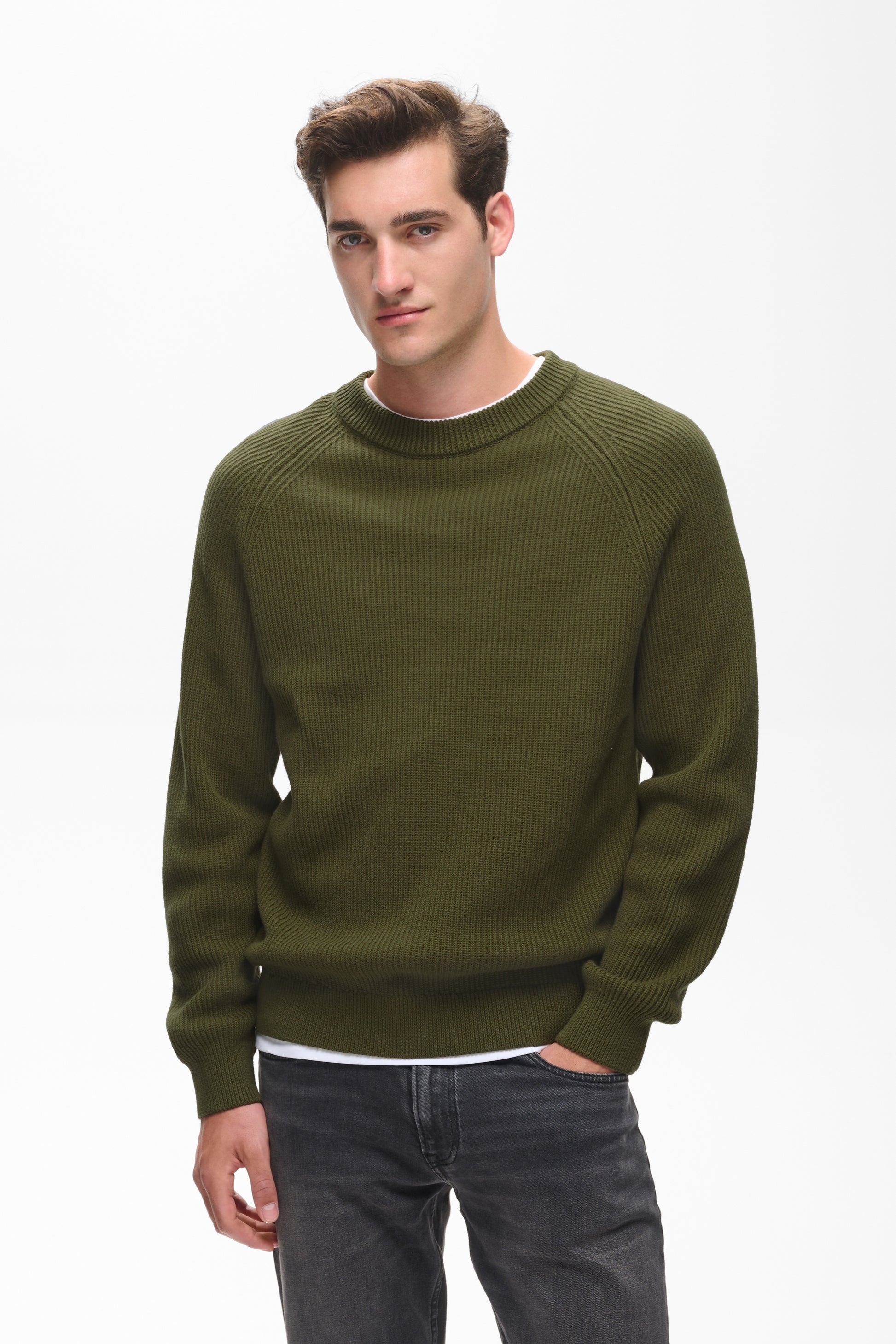 A man wearing an olive green ribbed knit crewneck sweater over a white undershirt and dark gray jeans, photographed against a plain white studio background.