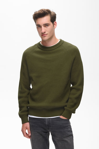 A man wearing an olive green ribbed knit crewneck sweater over a white undershirt and dark gray jeans, photographed against a plain white studio background.