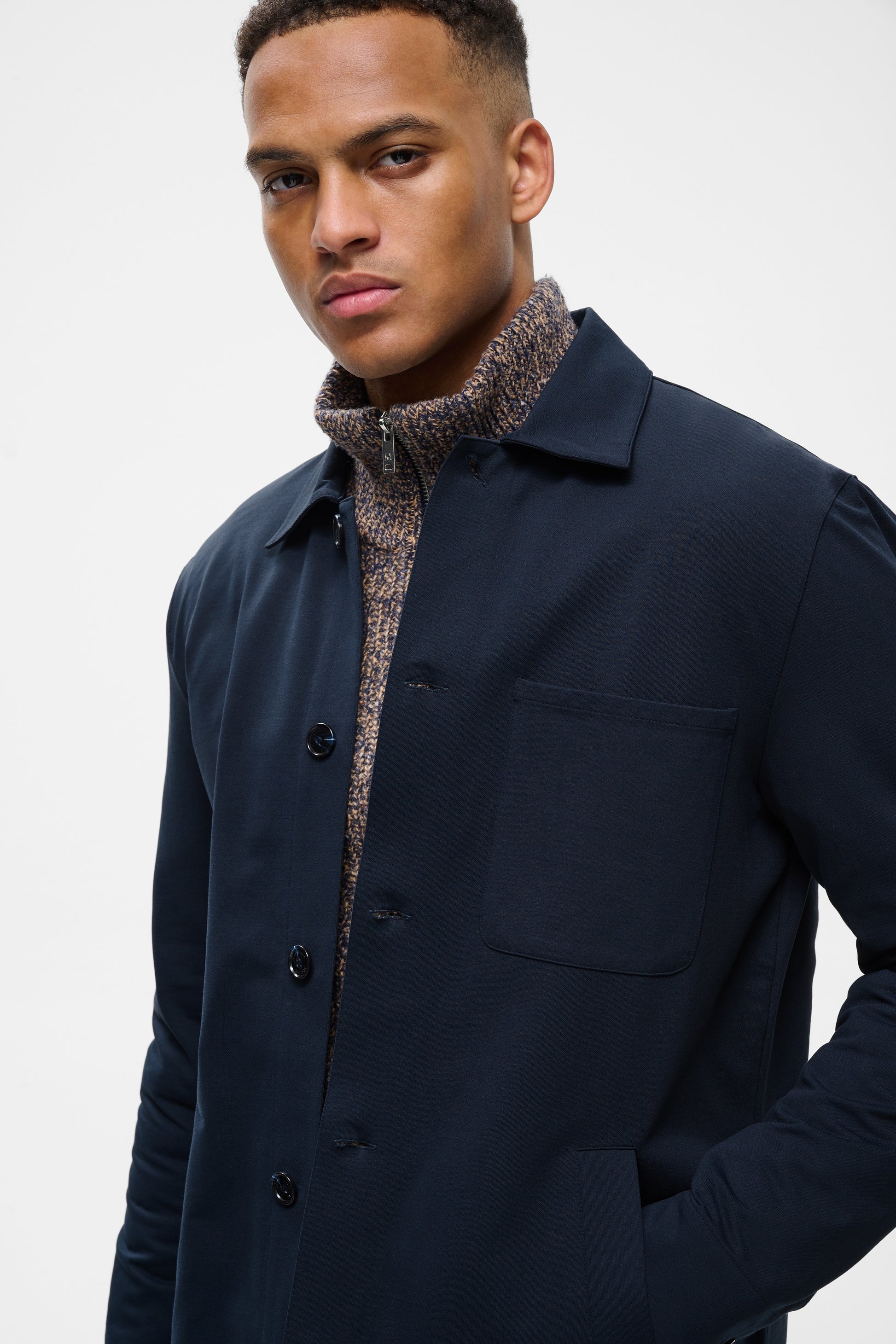 A man wearing a dark navy blue button-up overshirt over a textured brown and beige knit turtleneck sweater, photographed against a clean light gray studio background.