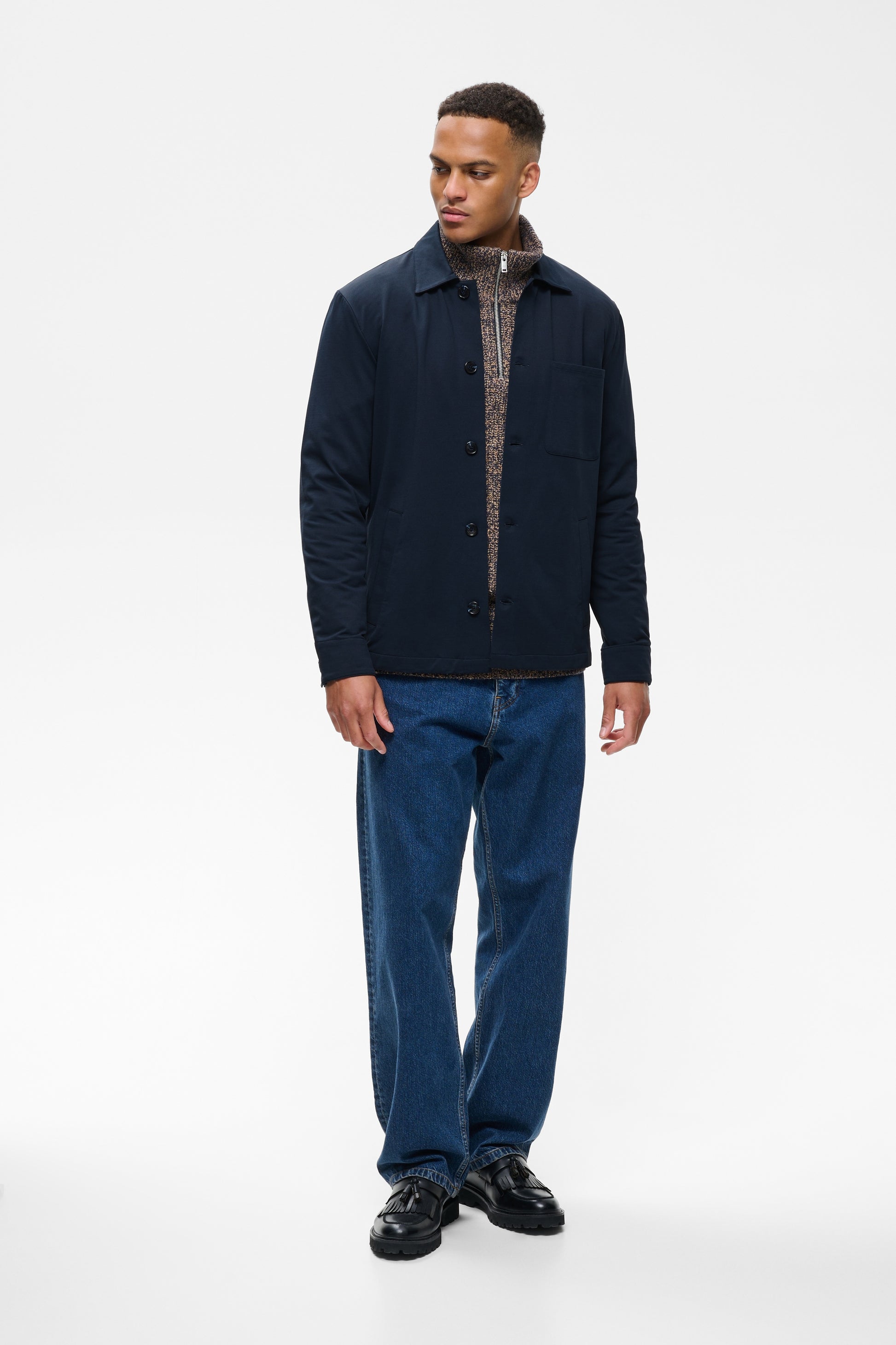 A man modeling a dark navy blue quilted jacket over a patterned shirt and blue jeans, standing against a plain white studio background.