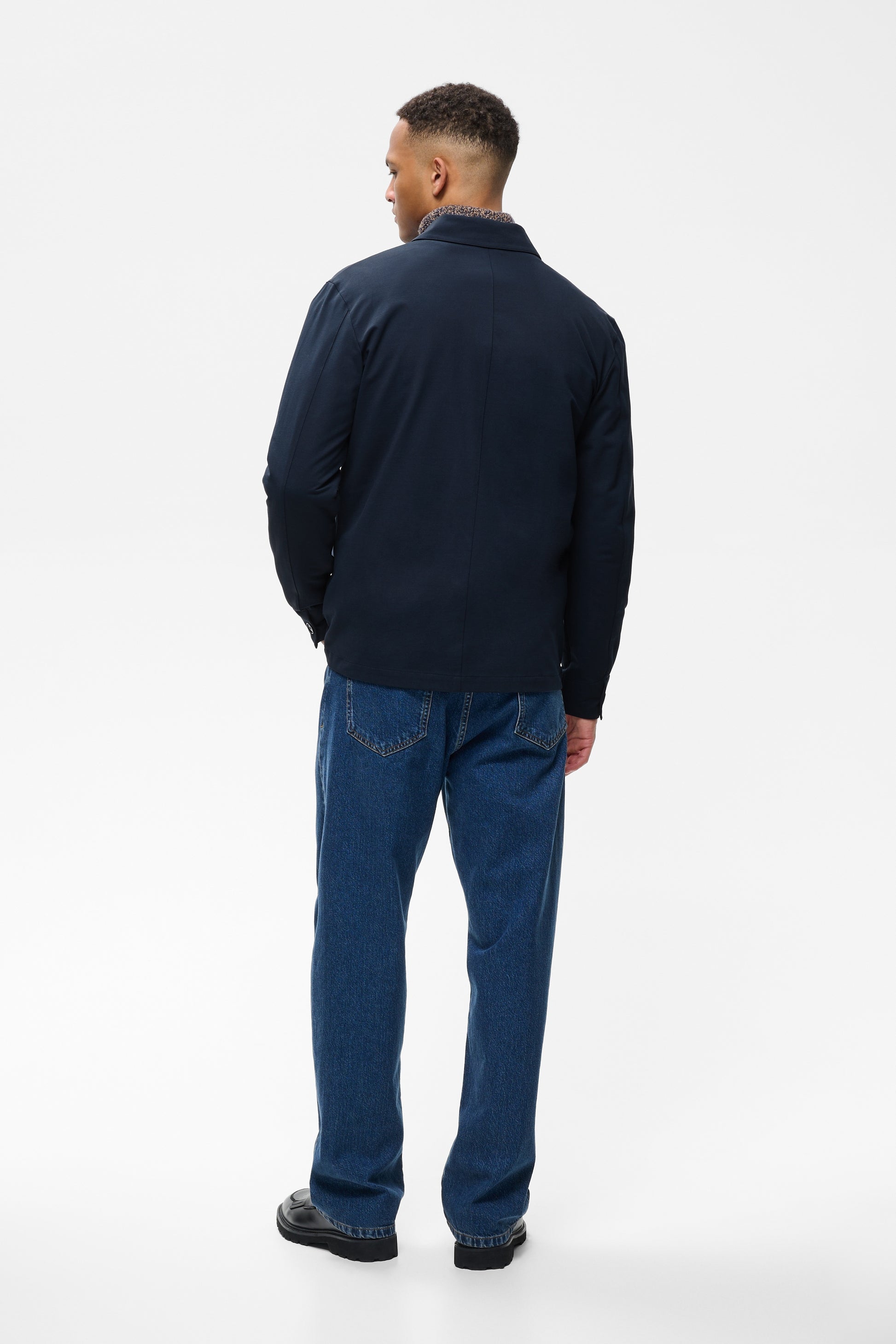 A man viewed from the back wearing a dark navy blue zip-up jacket and blue jeans, standing against a plain white studio background.