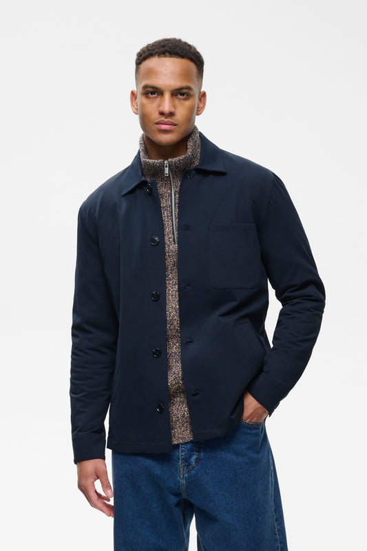 A man wearing a dark navy blue utility jacket over a patterned brown and beige shirt, paired with blue jeans, photographed against a plain light gray studio background.