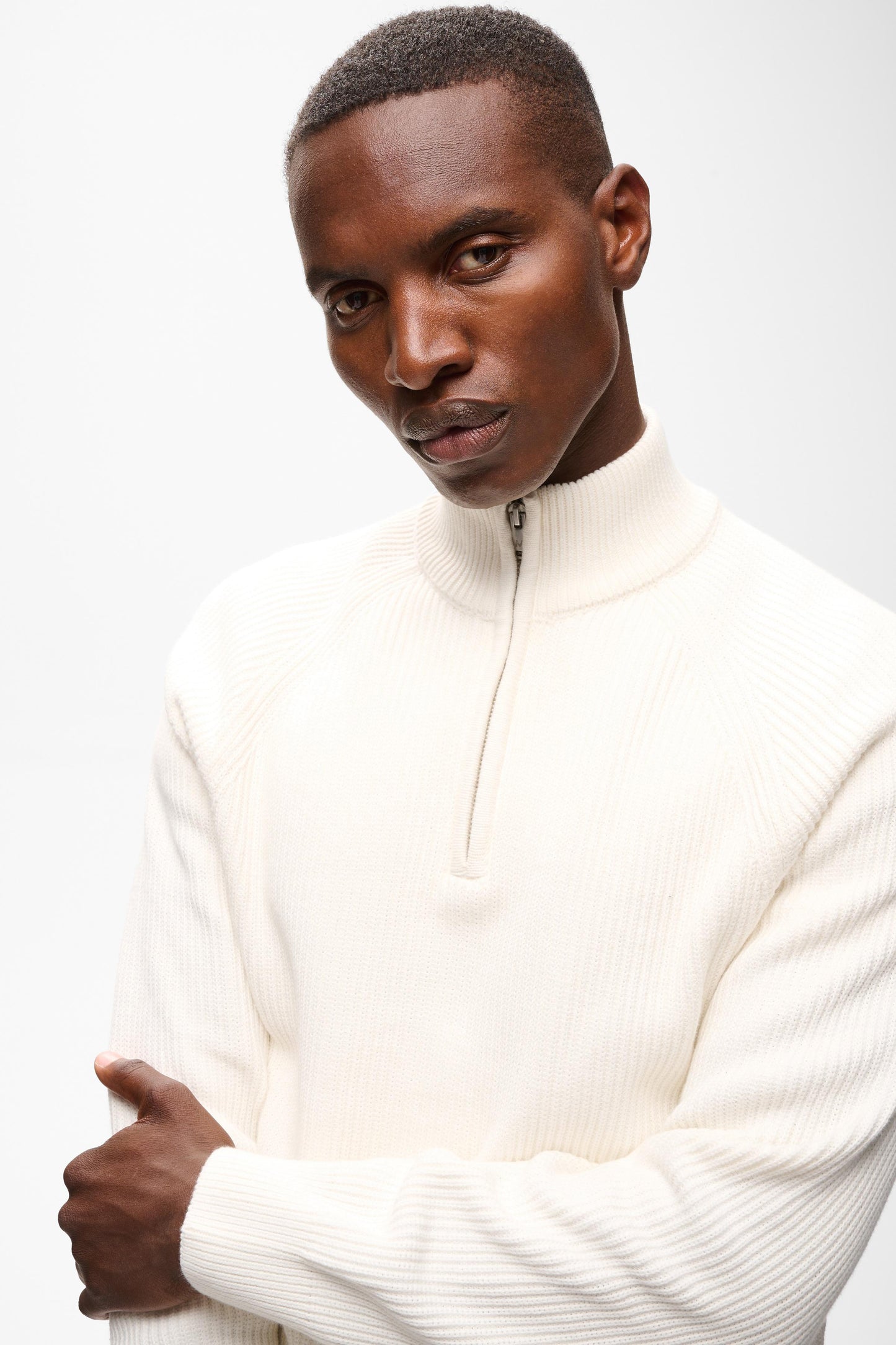 A man wearing a cream-colored ribbed knit mock-neck quarter-zip sweater, photographed against a clean white studio background.