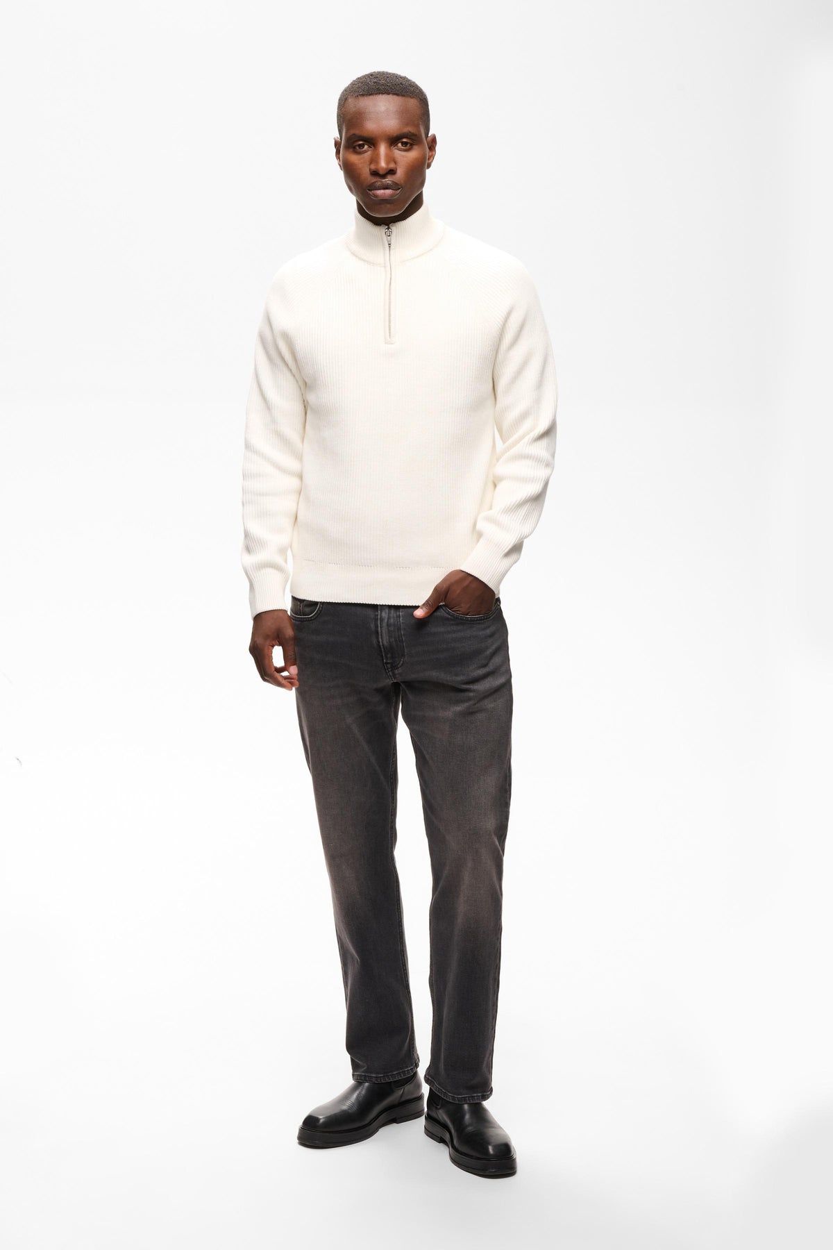 A man modeling a cream-colored quarter-zip knit sweater paired with dark gray jeans and black leather shoes, standing against a plain white studio background.