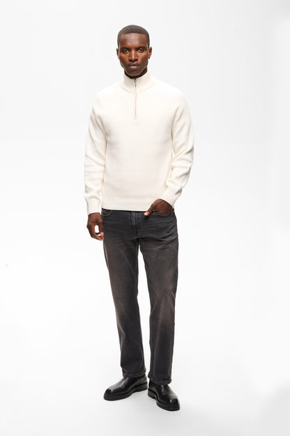 A man modeling a cream-colored quarter-zip knit sweater paired with dark gray jeans and black leather shoes, standing against a plain white studio background.