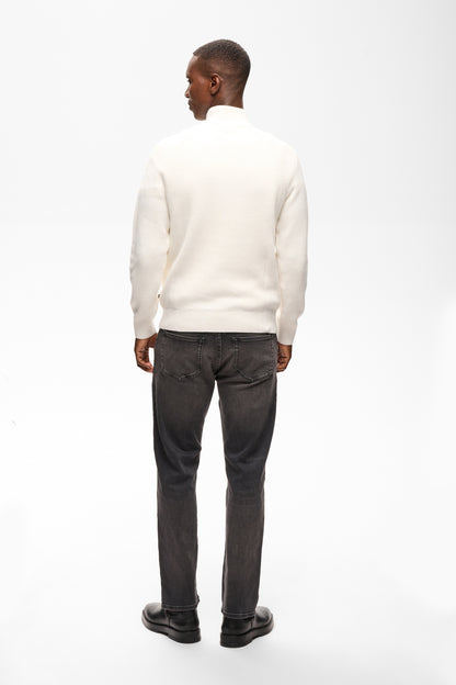 A man viewed from behind wearing a cream-colored mock neck sweater, dark gray jeans, and black shoes, standing against a plain white studio background.