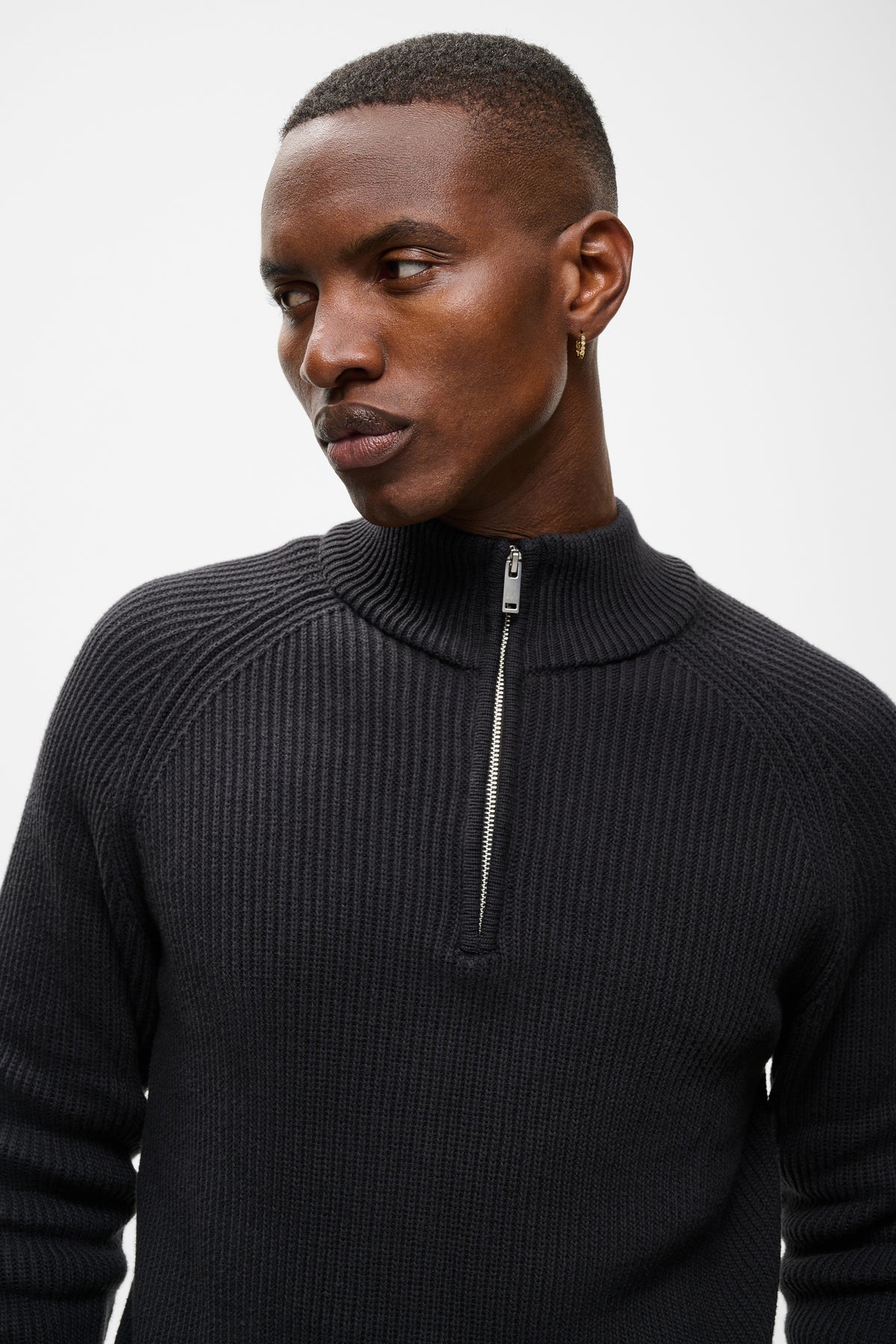 A man wearing a black ribbed quarter-zip mock neck sweater, shown from the chest up against a light gray studio background.