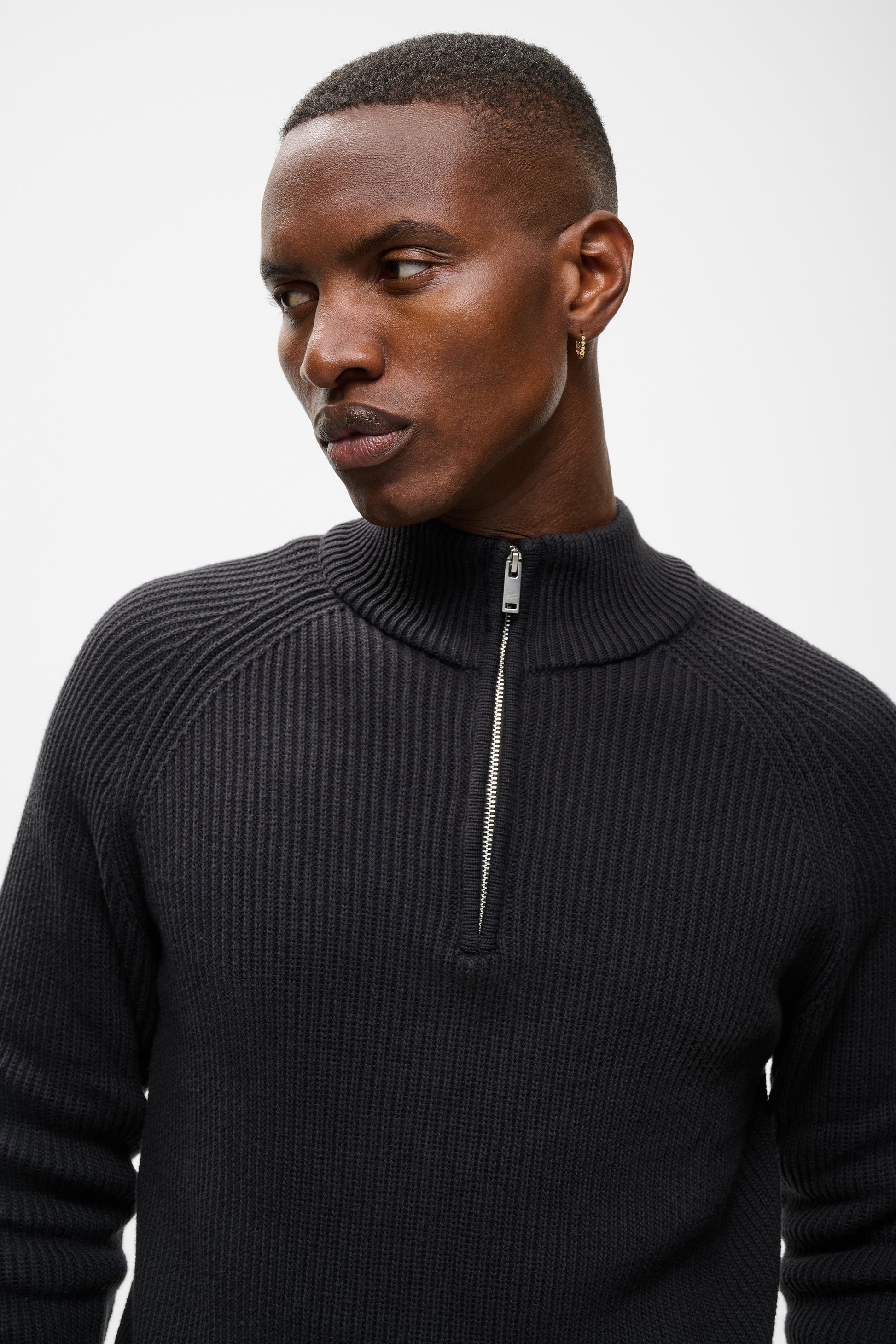A man wearing a black ribbed quarter-zip mock neck sweater, shown from the chest up against a light gray studio background.