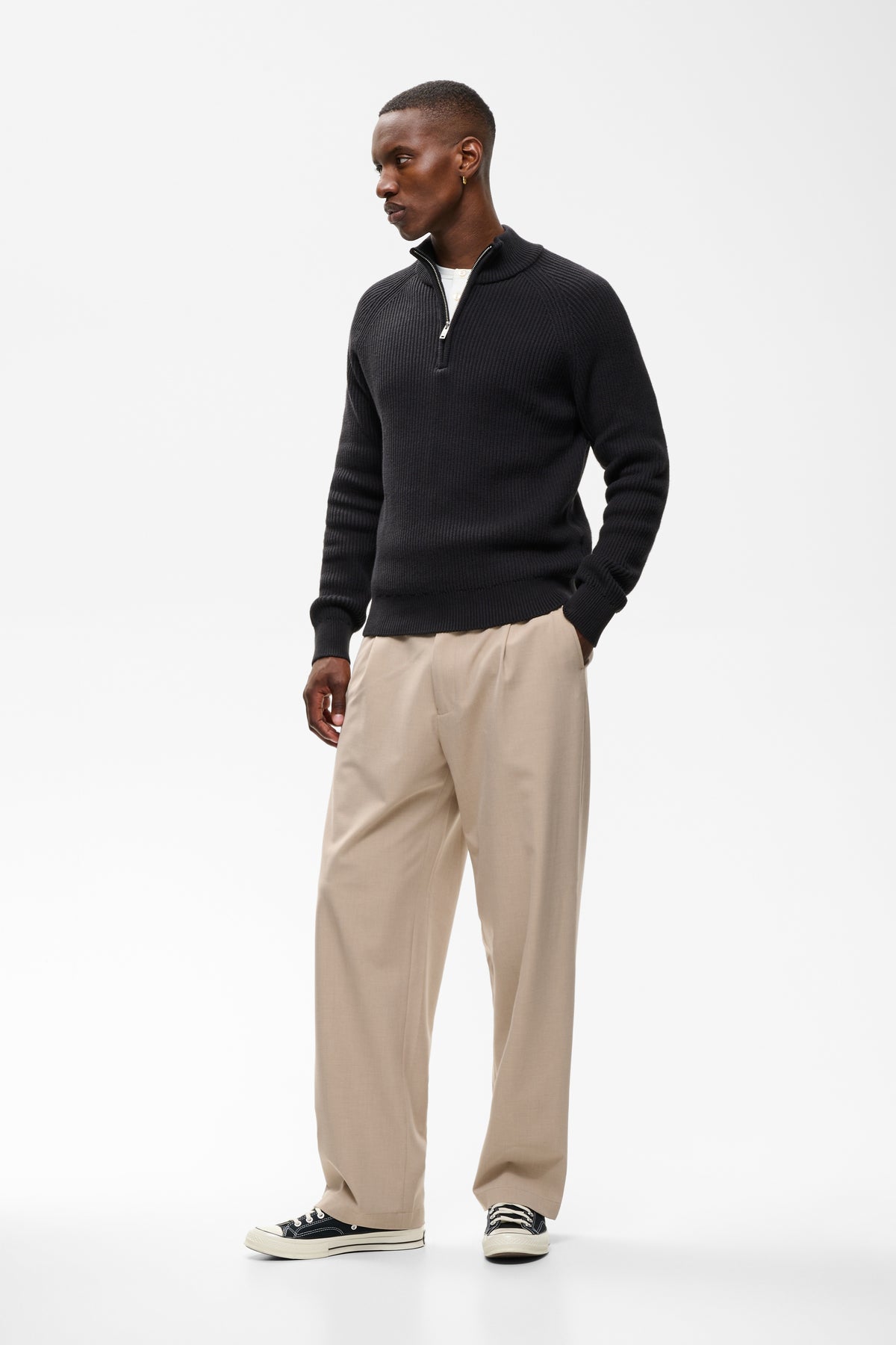 A man modeling a black long-sleeve quarter-zip pullover sweater, paired with beige trousers and black-and-white sneakers, against a plain white studio background.